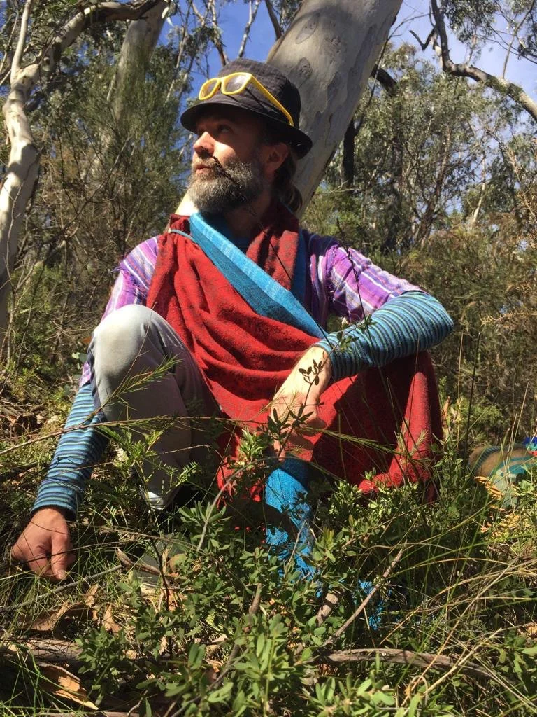 Person sitting in a eucalyptus forest wearing colorful layered clothing, yellow sunglasses, and a red scarf while looking thoughtfully into the distance.