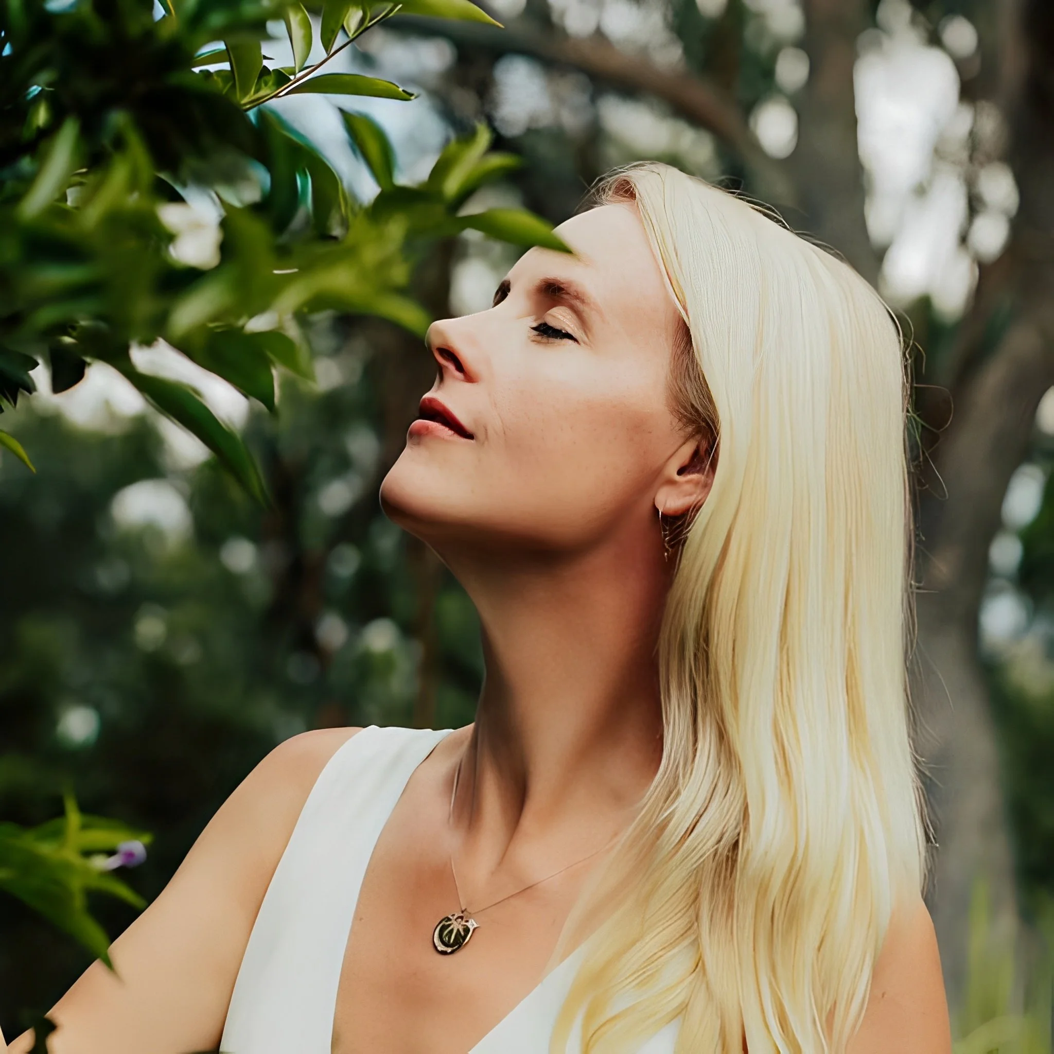 Close-up profile of a blonde woman outdoors with eyes closed near green leaves, appearing calm, reflective, and connected to nature.