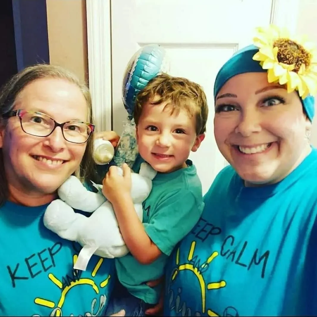 middle age woman in glasses holds grandson carrying a balloon and stuffed animal with smiling woman wears a headscarf with a sunflower on it