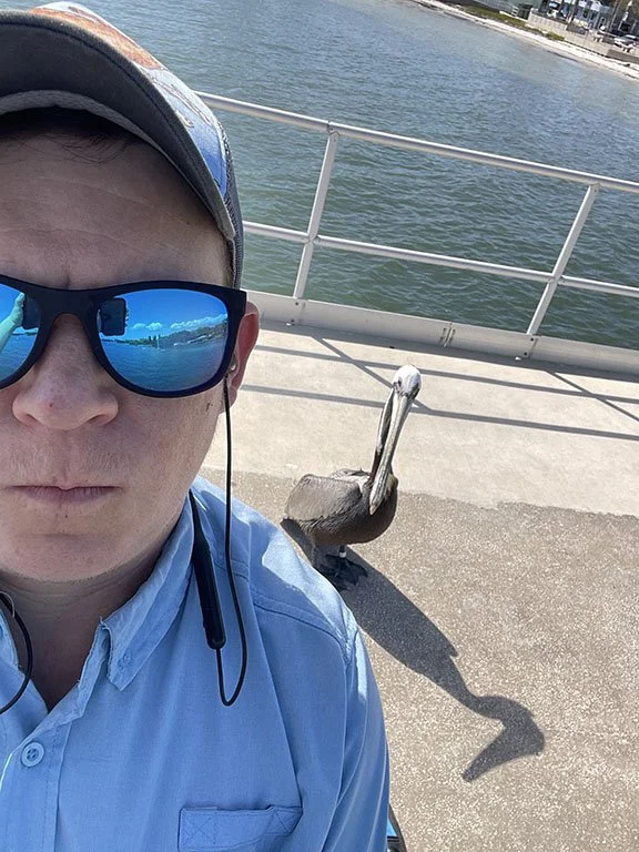 man in a blue button up shirt and reflective blue sunglasses stands in front of a pelican along a pier on a body of water
