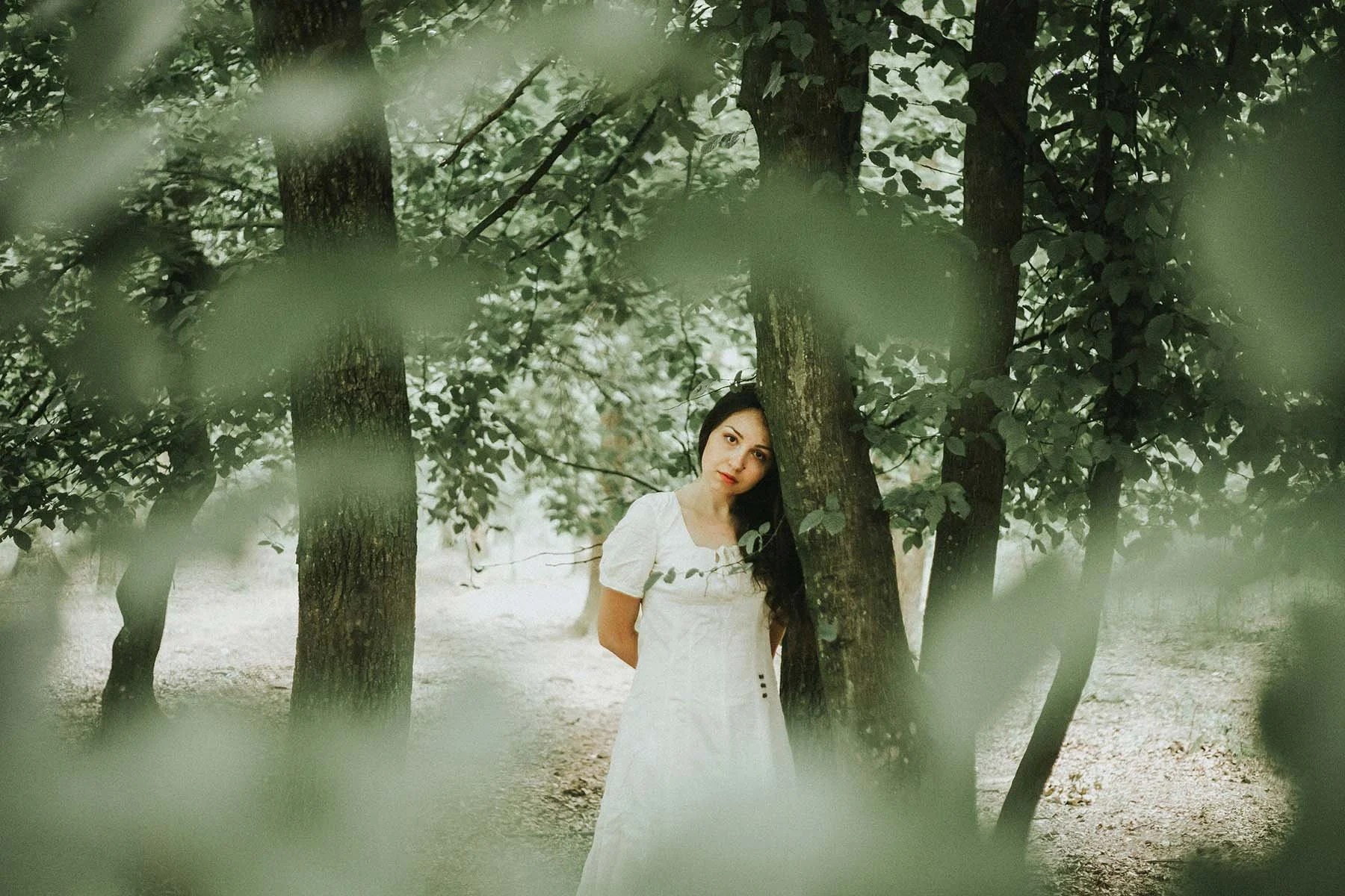 Woman in a white dress standing among trees in a forest, partially framed by out-of-focus leaves in the foreground.