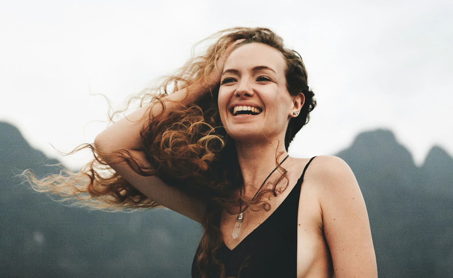 A smiling person with long curly hair stands outdoors with mountains in the background, laughing as the wind blows through their hair.
