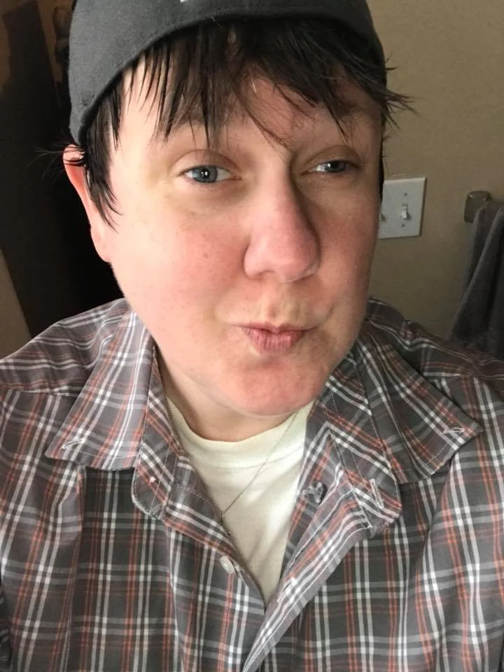 A person wearing a gray baseball cap, plaid button-up shirt, and white T-shirt looks slightly to the side while posing indoors.