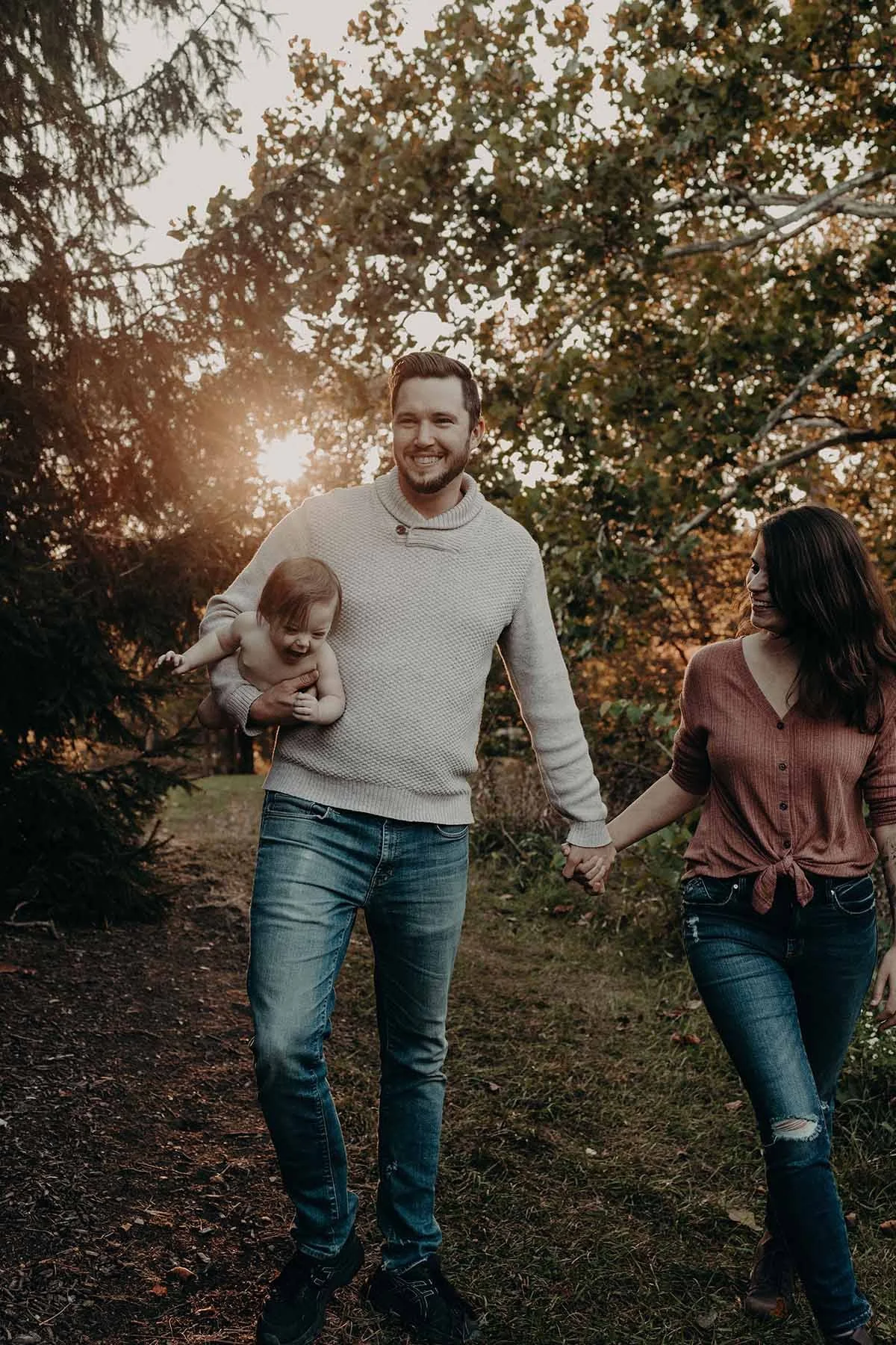 Family walking through a wooded area at sunset while holding hands and carrying a young child.