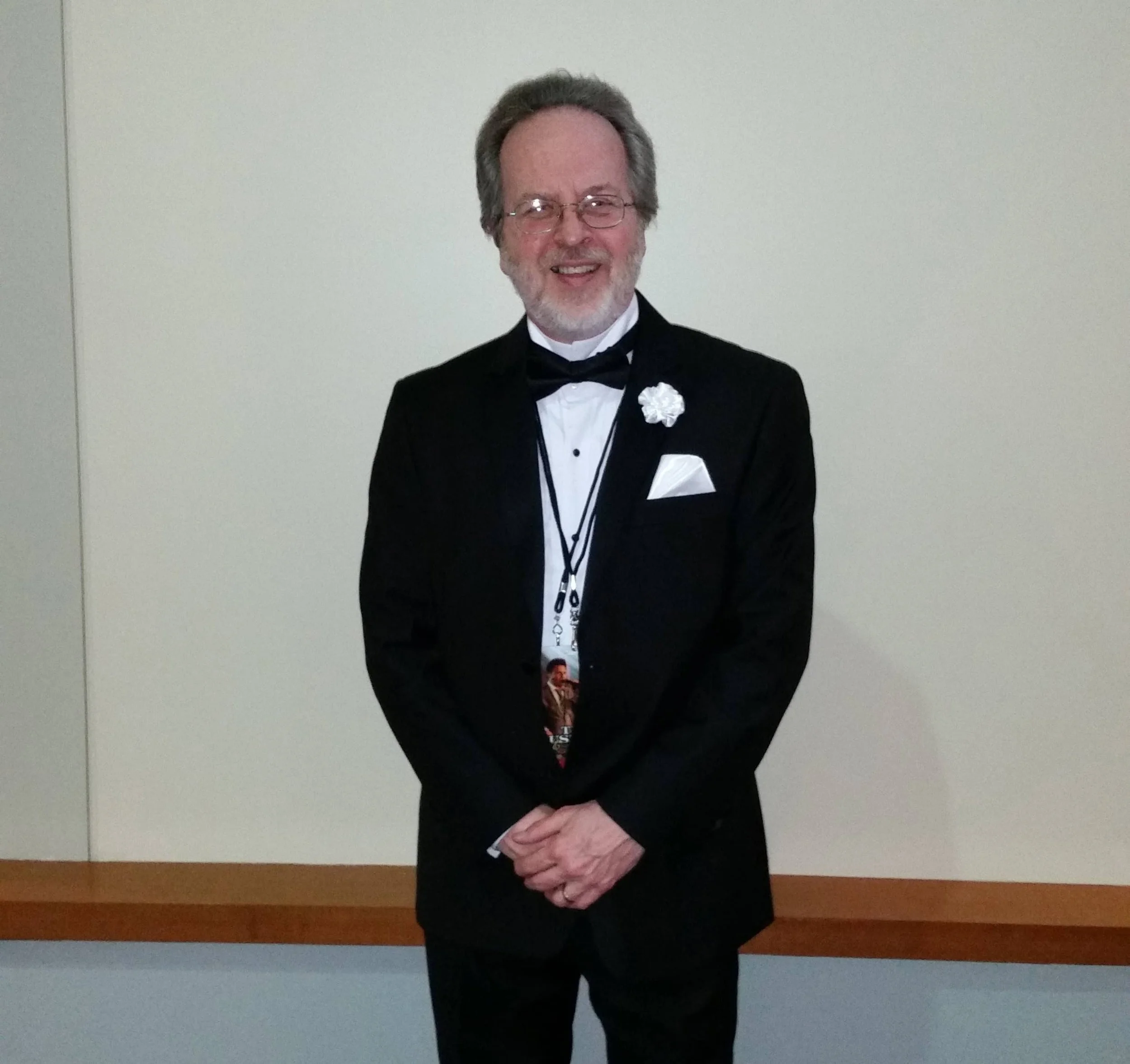 smiling man with gray hair and white beard wears a black tuxedo and glasses