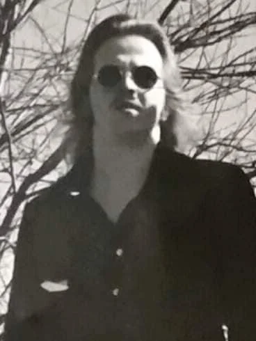 black and white photo of a man with long hair standing in front of tree branches with round sunglasses on