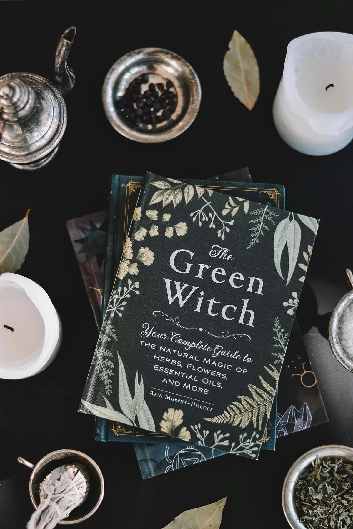 Flat-lay photograph of The Green Witch book surrounded by candles, dried herbs, bowls, and ritual-inspired objects arranged on a dark surface.