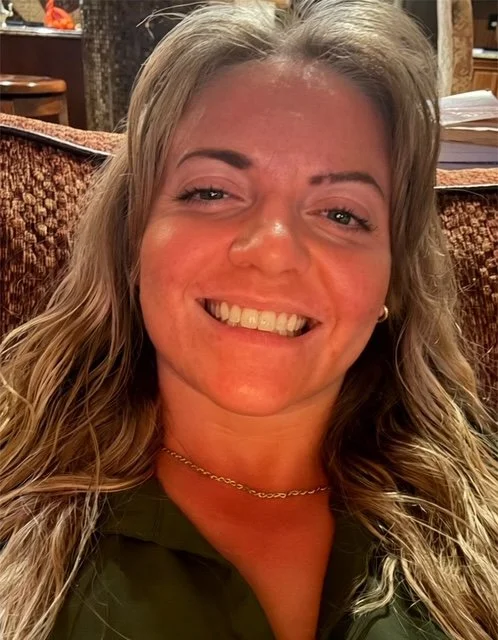 Close-up portrait of a smiling blonde woman with long, wavy hair, wearing a dark green top and gold necklace while seated indoors against a warm brown background.
