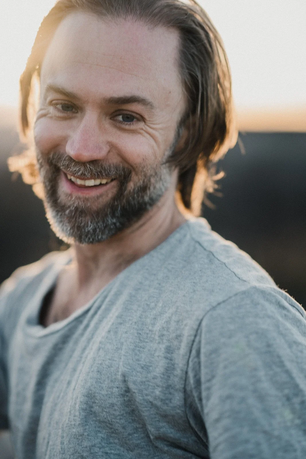 Close-up portrait of a smiling man outdoors in warm backlit sunlight with long hair and a gray shirt against a blurred natural background.