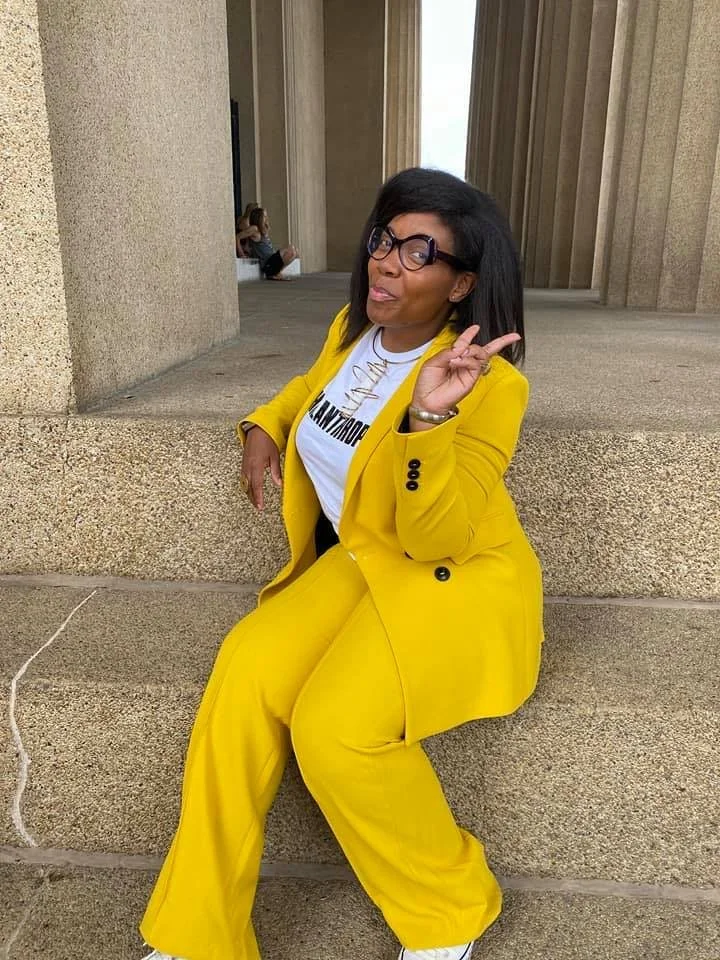 Woman in bright yellow suit and graphic T-shirt posing playfully between large concrete columns in an urban setting.
