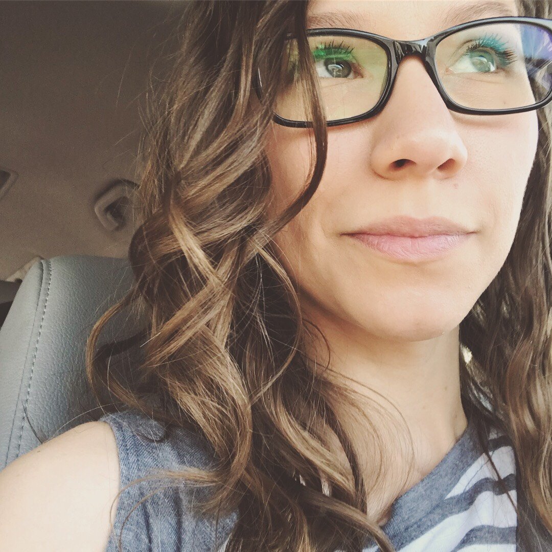 Portrait of a woman with long brown hair and glasses, smiling softly while seated in a car