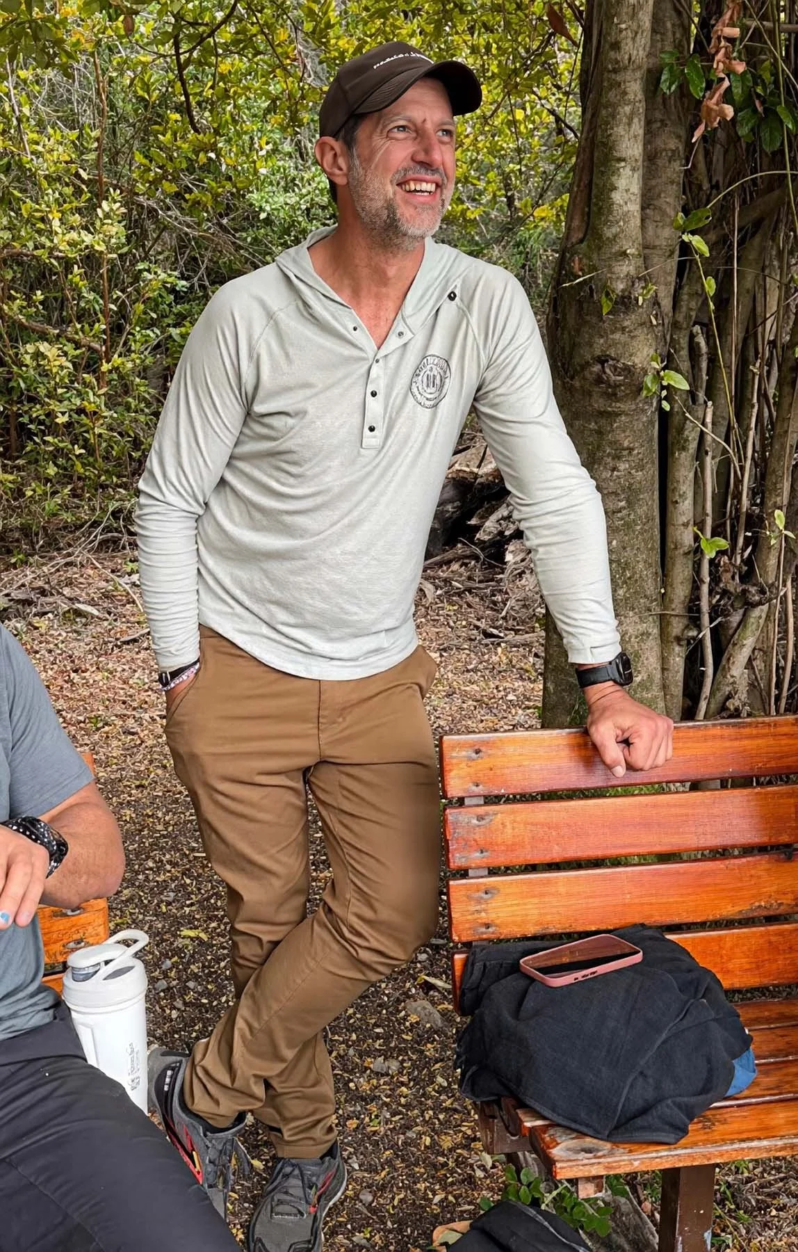 A smiling man in a black baseball cap, light gray long-sleeve shirt, and brown pants leans against a wooden bench in a leafy forest setting. A phone and folded jacket rest on the bench, and part of another seated person is visible at the left edge.