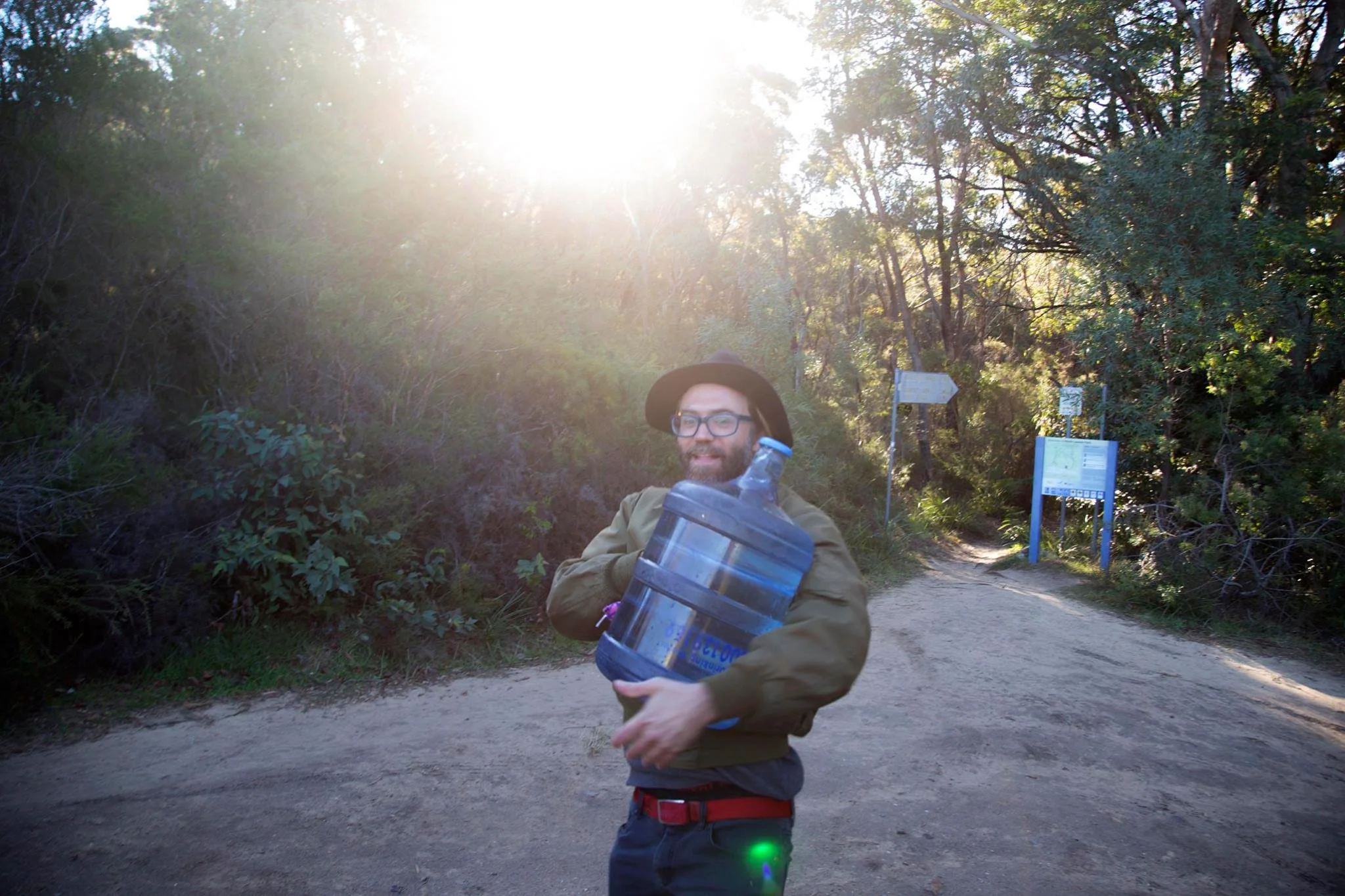 Person carrying a large water jug on a forest trail in the Blue Mountains, surrounded by eucalyptus trees and warm sunlight.