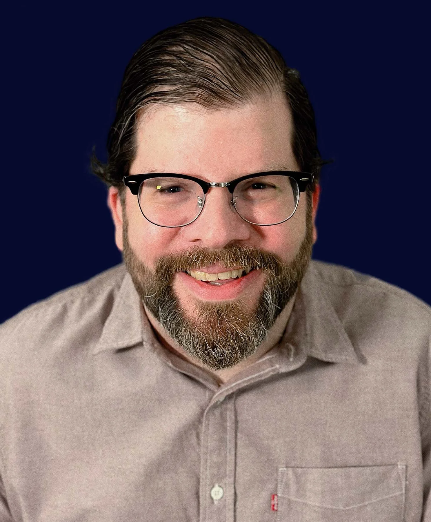 A smiling bearded man wearing glasses and a light gray button-up shirt poses against a solid dark navy background.