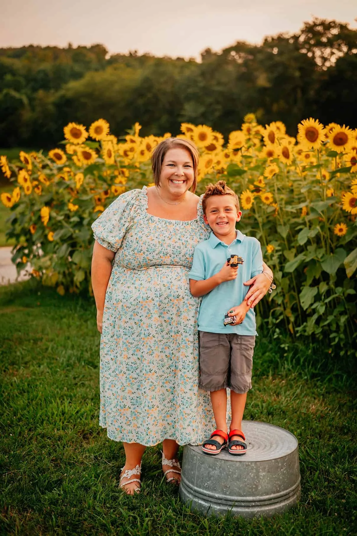 smiling woman stands in a sunflower field in a floral dress stands with young son wearing a blue polo shirt, shorts, and holding toy trucks in his hand