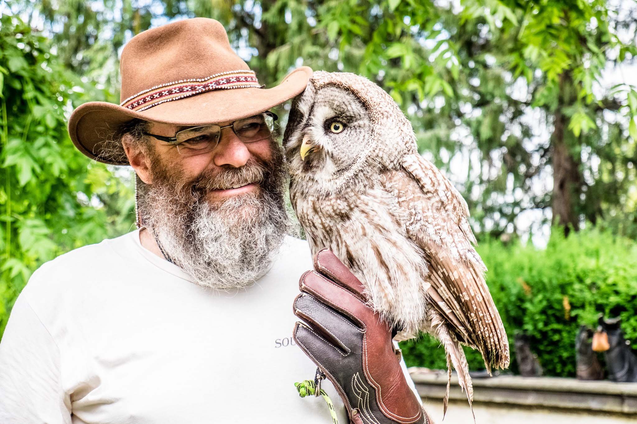 A smiling bearded man in a brown cowboy hat and glasses holds a large gray owl on a leather-gloved hand outdoors, surrounded by bright green trees and foliage.