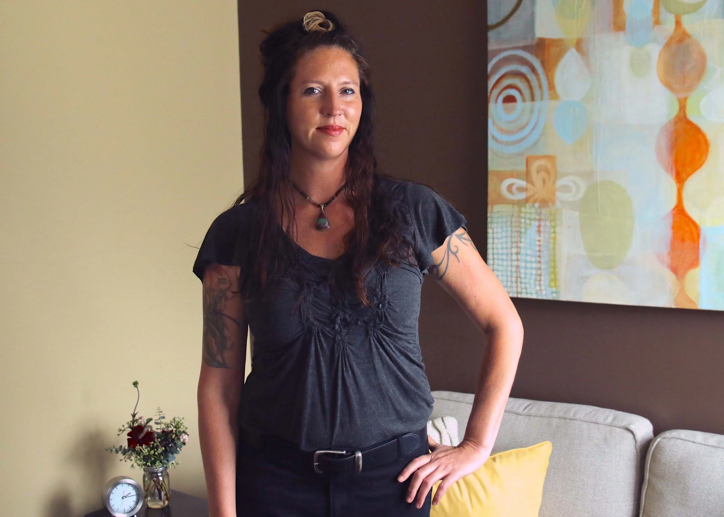 Woman with long dark hair standing indoors with hand on hip in a cozy living room.