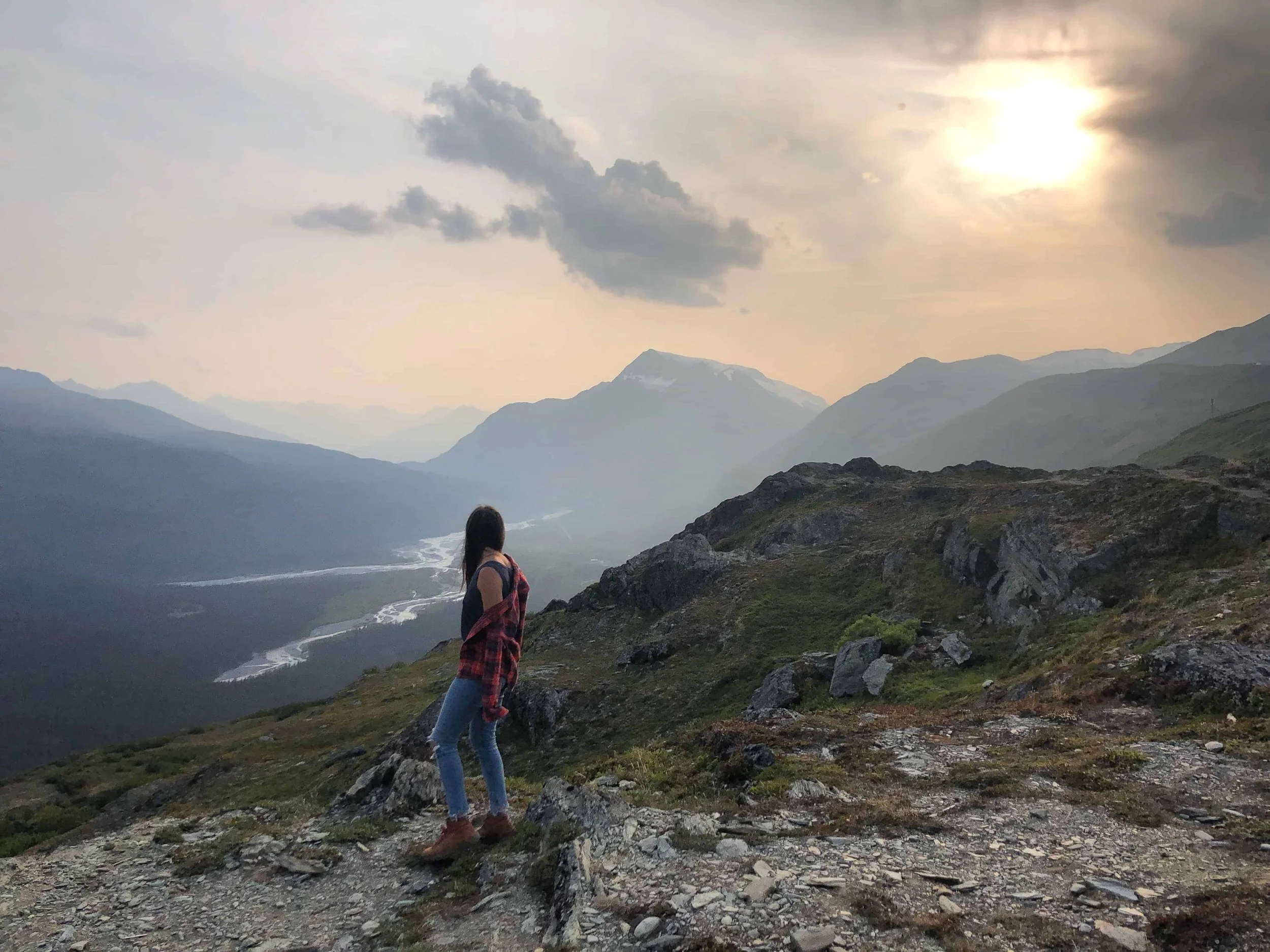 Woman standing on a mountain facing away toward the horizon with the sun glowing behind the clouds.