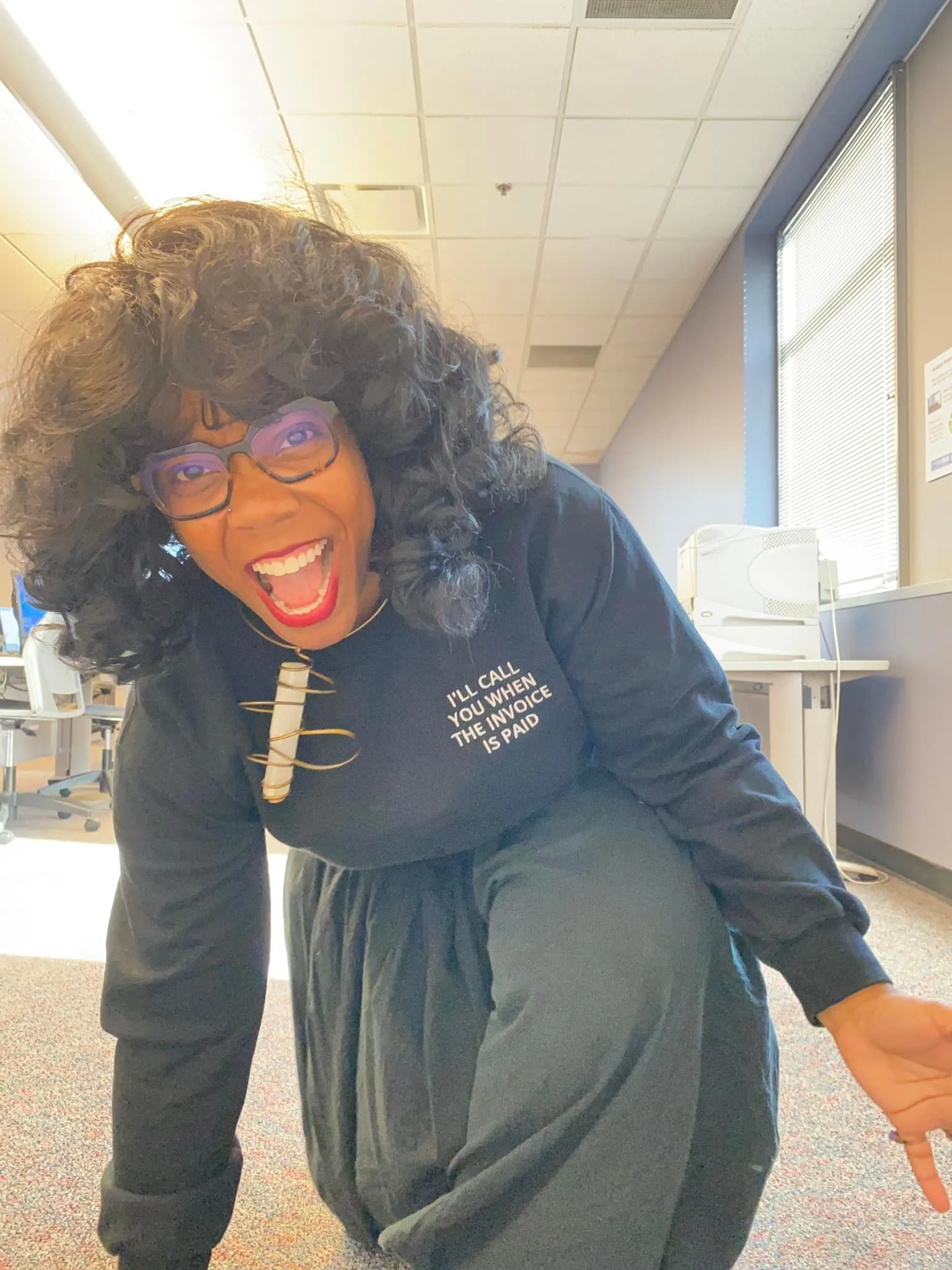 Woman laughing in an office hallway wearing glasses and a black outfit with a shirt reading “I’ll call you when the invoice is paid.”