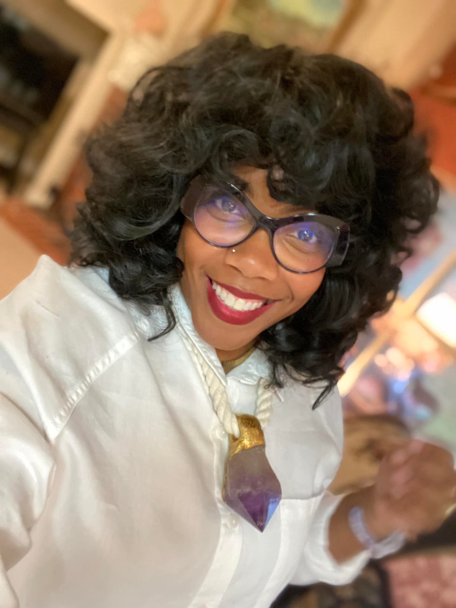 Smiling woman with curly black hair, glasses, red lipstick, and crystal necklace wearing a white button-up shirt indoors.