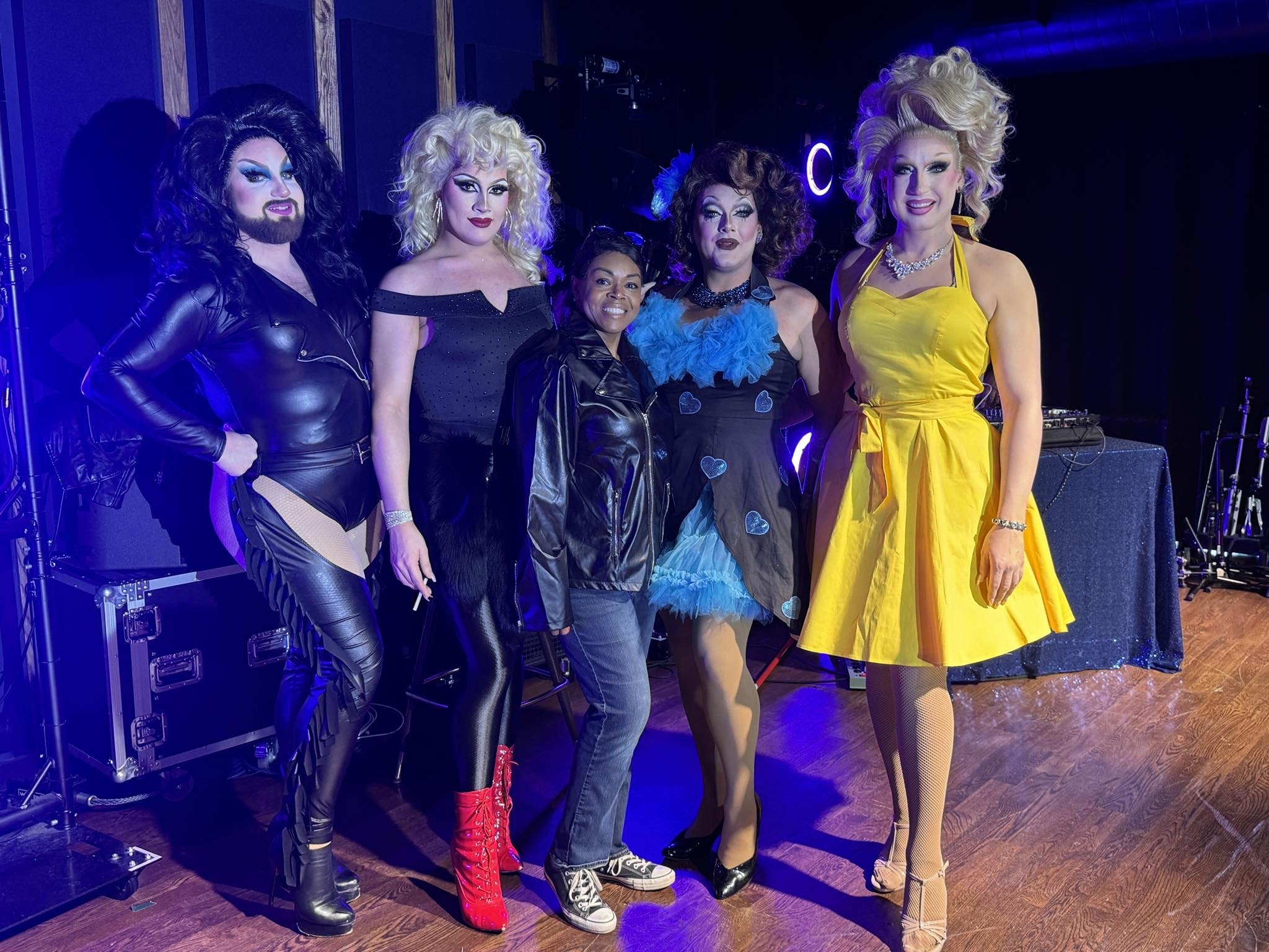 Four drag queens pose on stage with Black woman wearing a leather jacket, jeans, and Converse sneakers