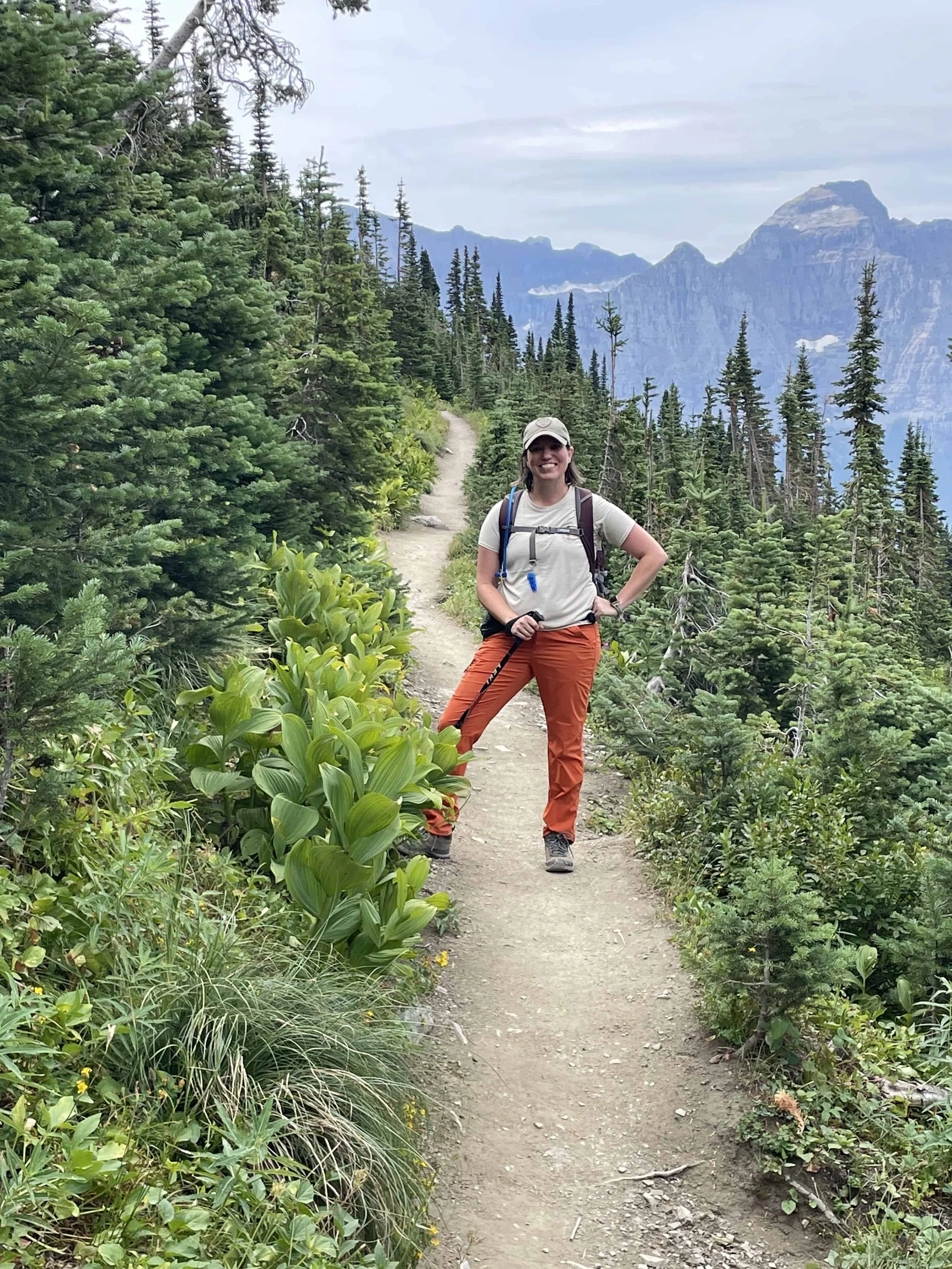 Woman in hat and orange hiking pants smiling on a mountain trail with hand on hip.