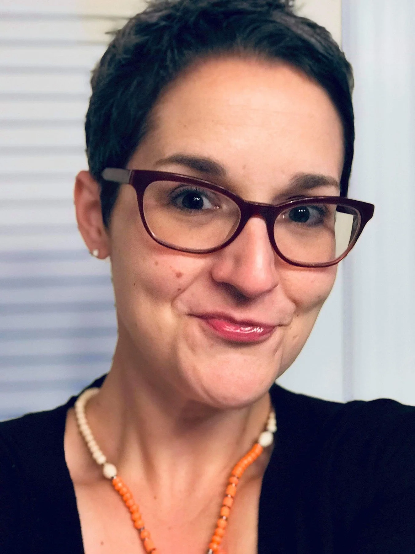 A person with short dark hair and glasses smiles at the camera while wearing a black shirt and a beaded necklace indoors.