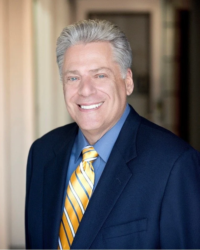 Professional headshot of a smiling man in a dark suit with a blue shirt and striped yellow tie.