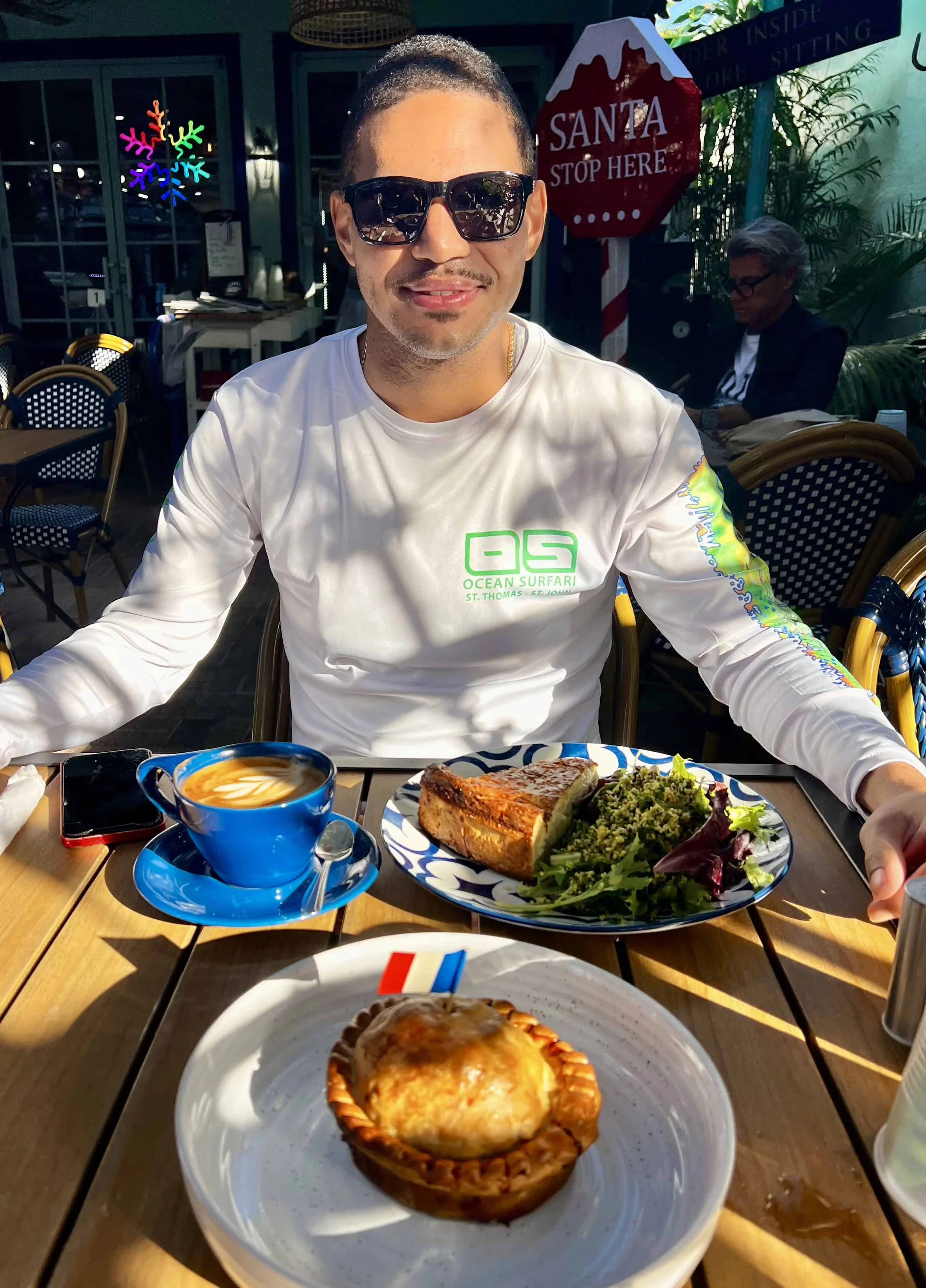 Ty Briggs smiles at an outdoor café table wearing sunglasses and a white long-sleeve shirt, seated in front of plates of food and coffee in bright natural sunlight.