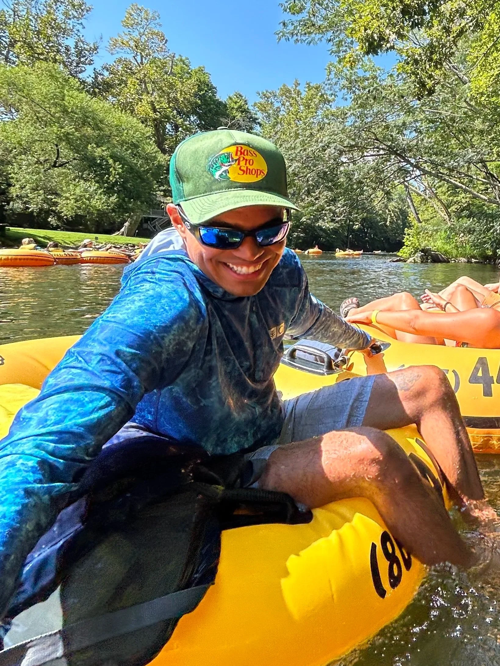 Ty Briggs smiles while floating down a river in a yellow inner tube, wearing sunglasses and outdoor clothing with trees, blue sky, and other tubers visible in the background.