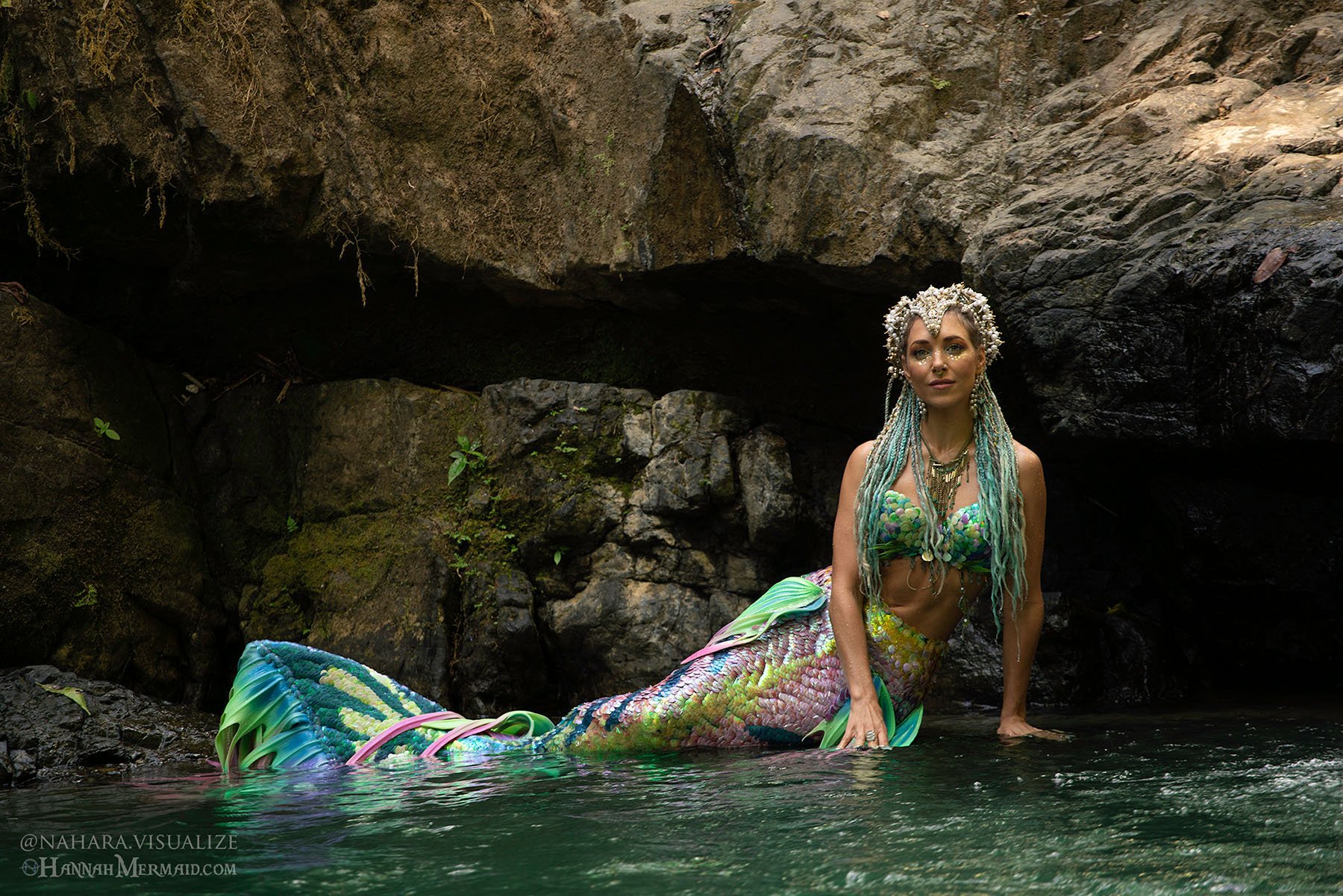 Woman in a colorful mermaid tail posing in a rocky jungle pool surrounded by dark volcanic stone and flowing water.