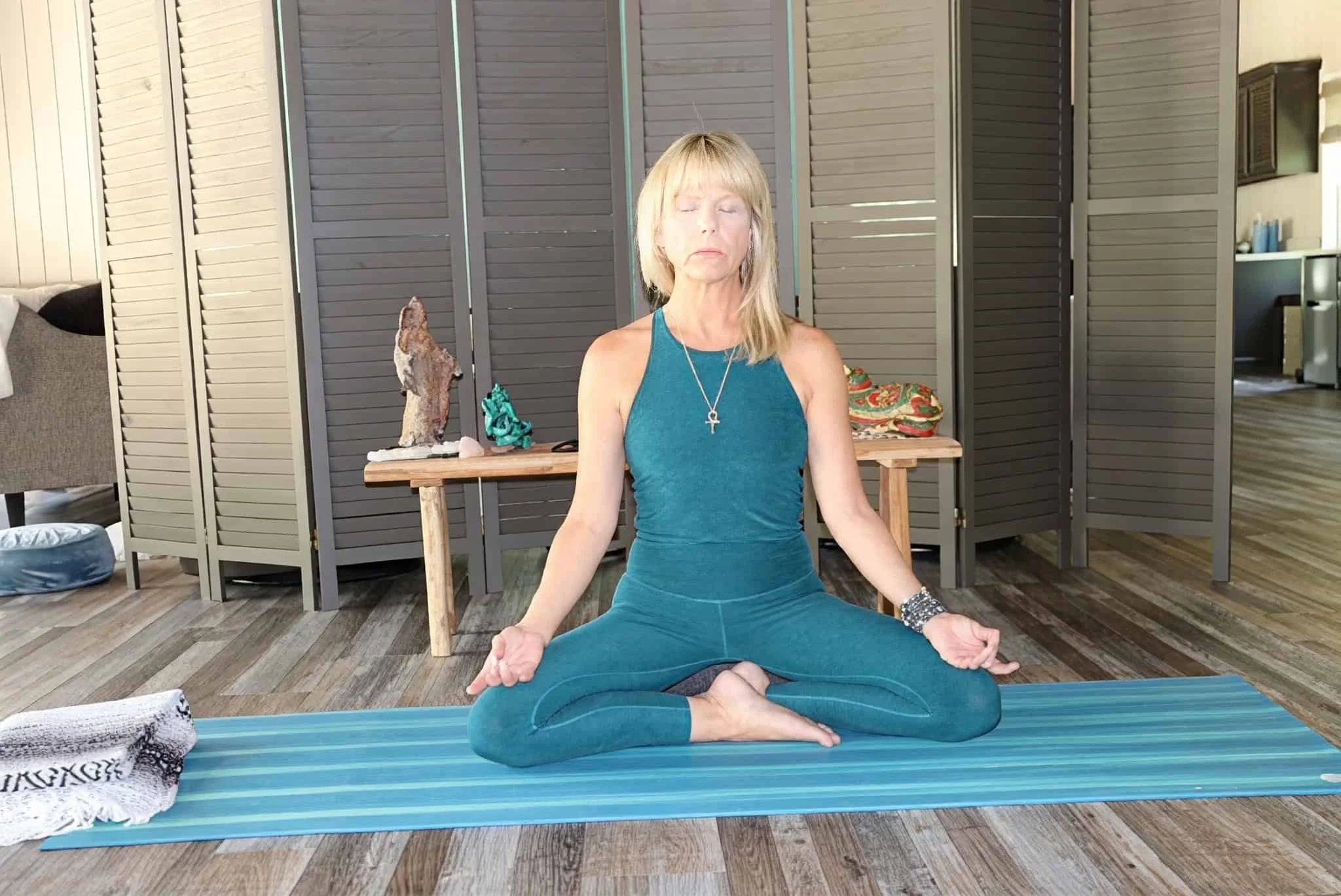 A woman sits in a cross-legged meditation pose on a yoga mat indoors, eyes closed, wearing a teal outfit in a calm, softly lit room.