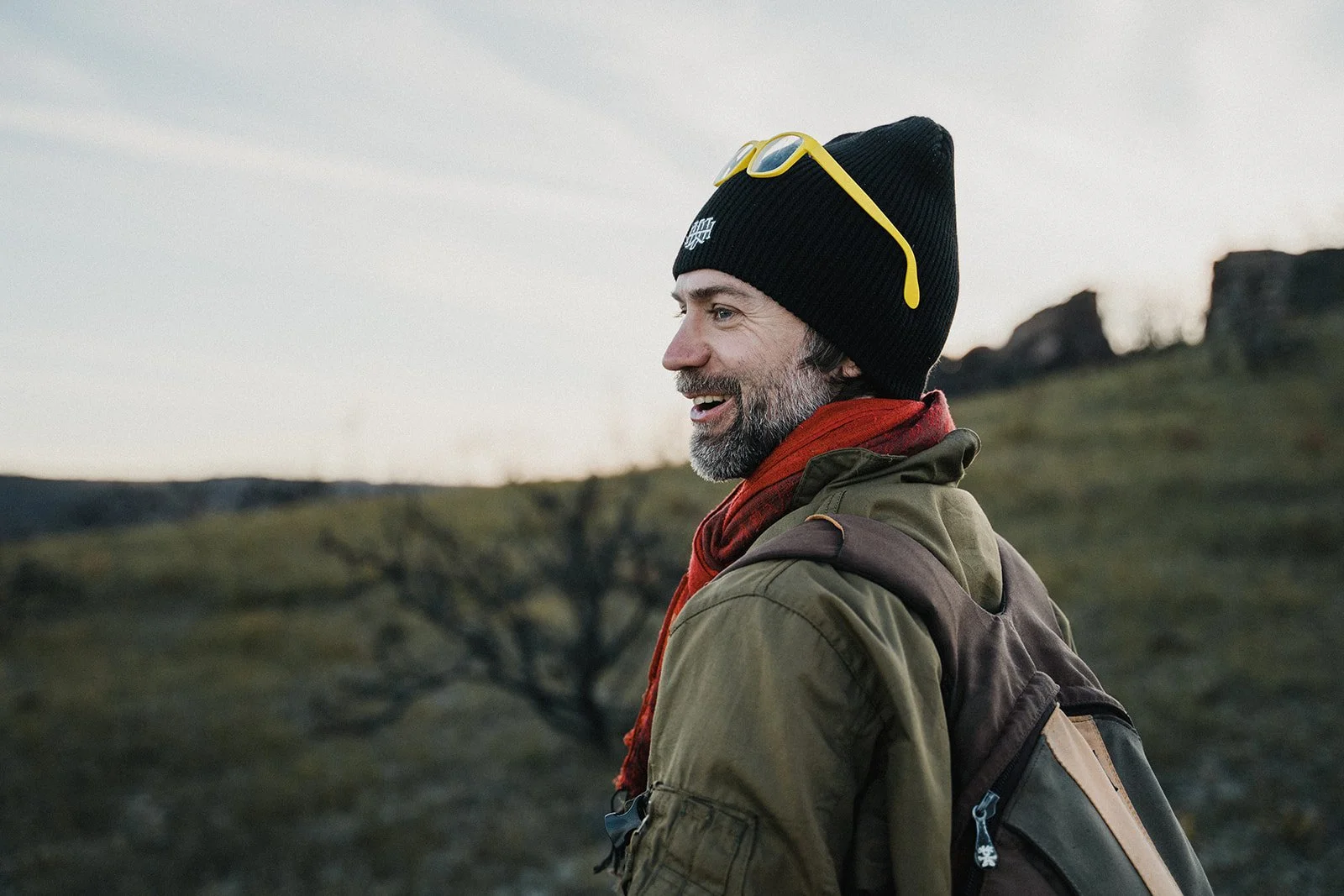 Smiling hiker wearing a backpack, red scarf, and black beanie standing outdoors in a rocky natural landscape at sunset.