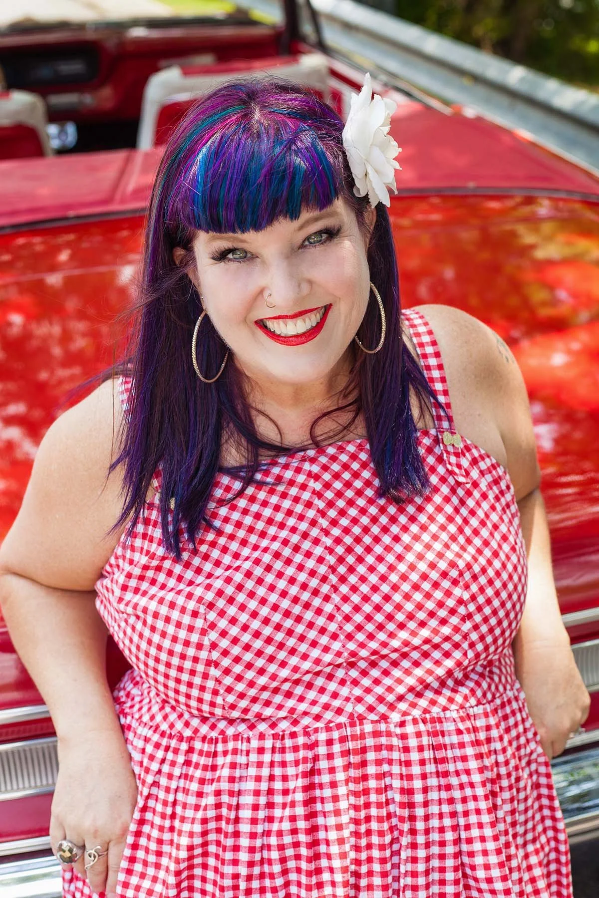 Portrait of a psychic medium with colorful purple and blue hair smiling in front of a vintage red convertible.