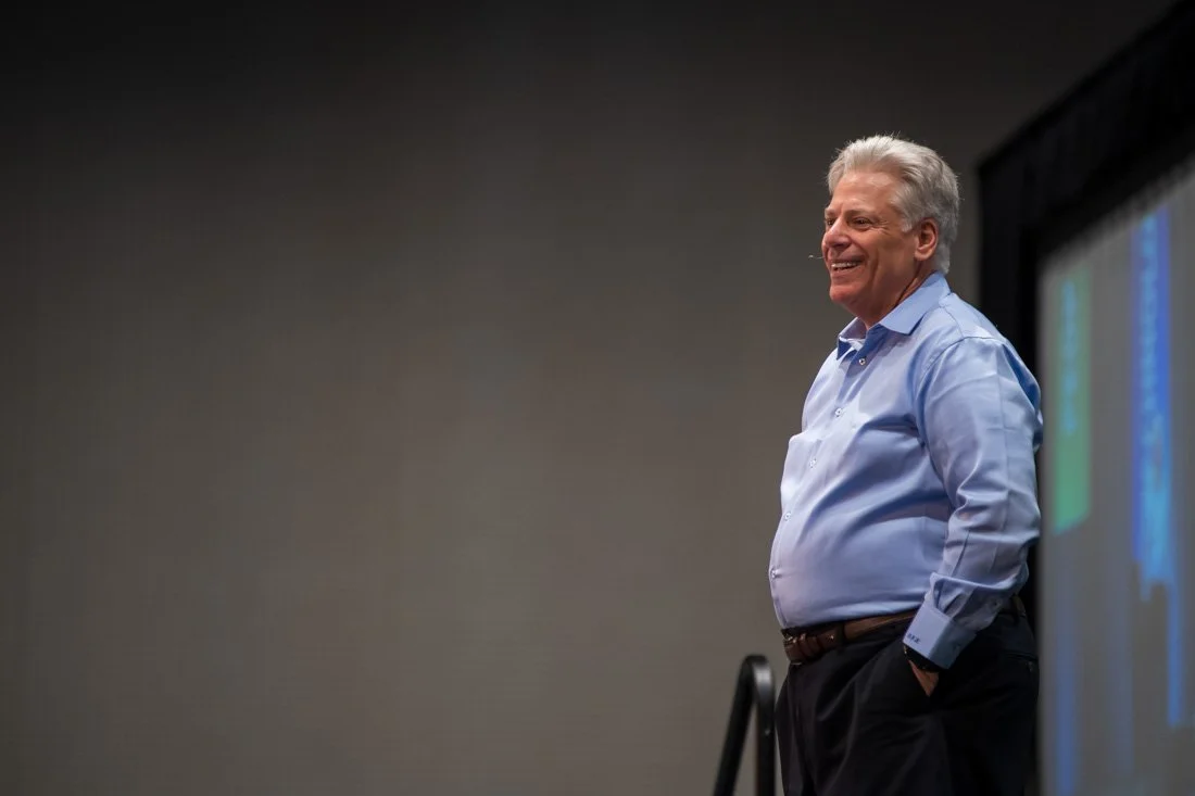 Man speaking onstage during a presentation while wearing a light blue dress shirt.