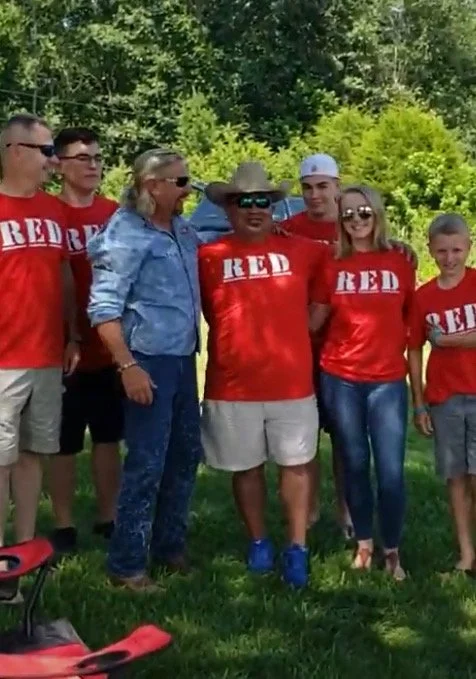 Group of adults and children wearing matching red shirts standing together outdoors on grass during a community event supporting veterans and military families.