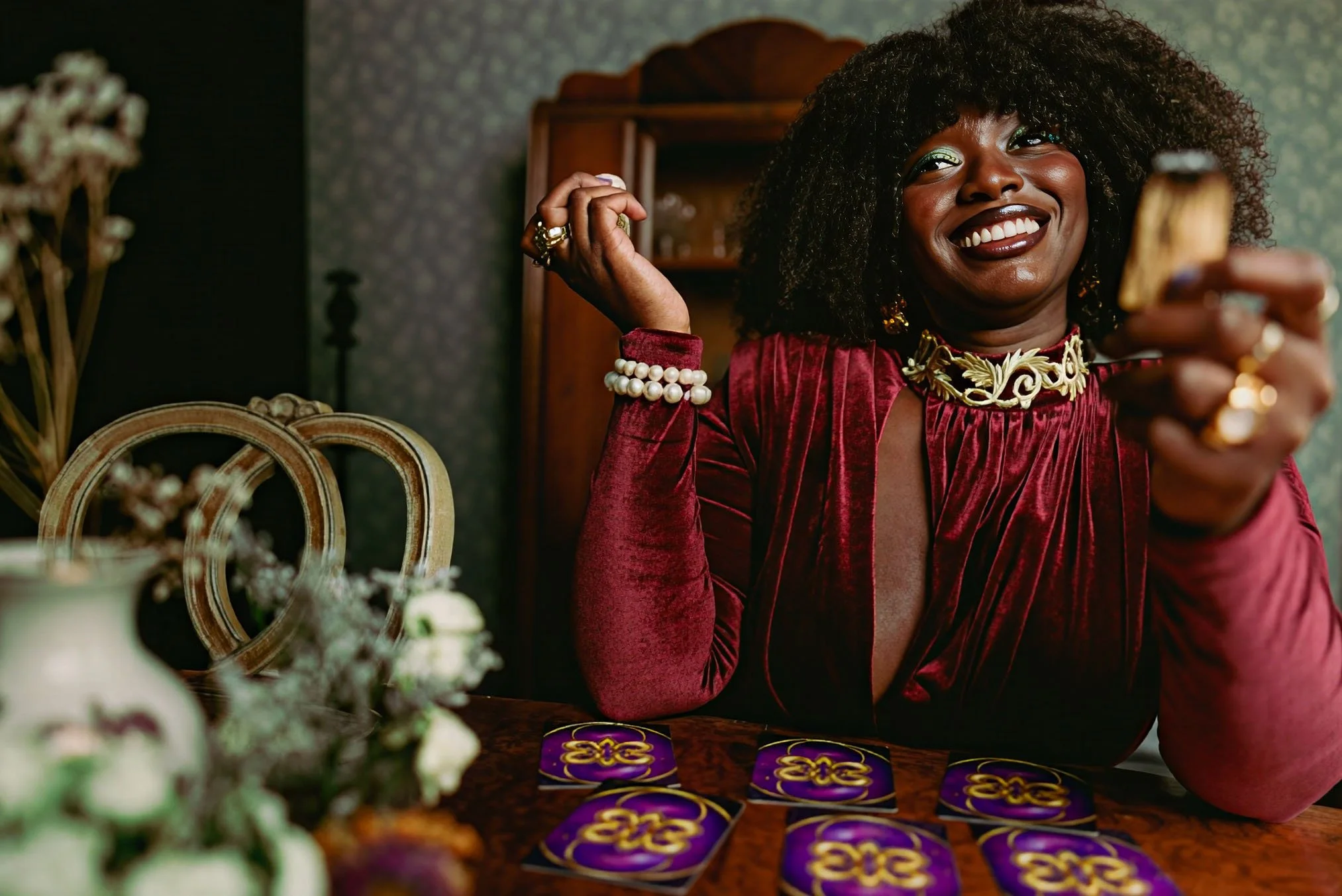 Delanyo Mensah smiles while holding a small piece of smoking wood at a table. She wears a deep red velvet outfit and gold jewelry, with purple and gold cards arranged in front of her and a decorative mirror and flowers in the background.