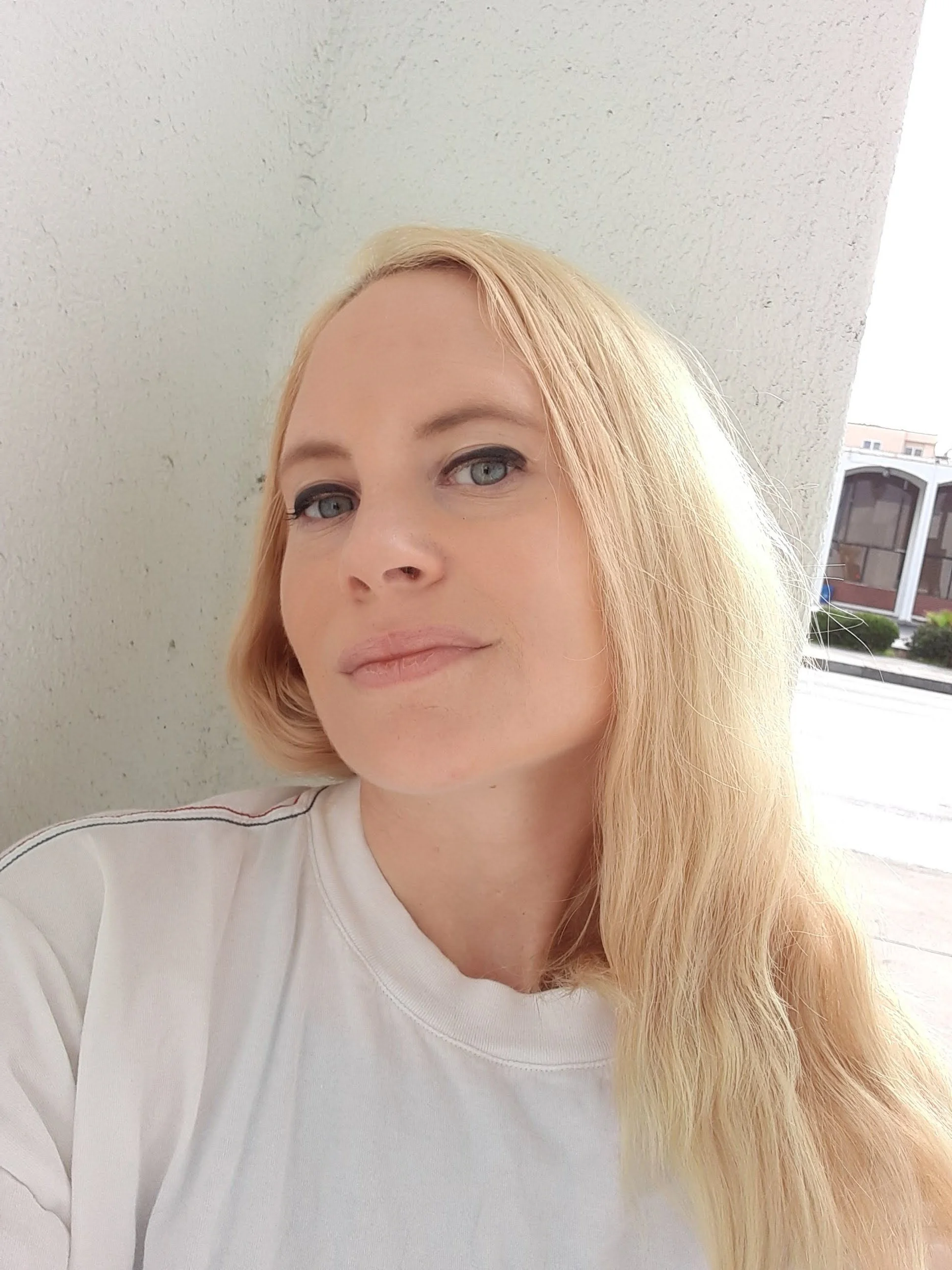 Selfie of a blonde haired woman outside against a white concrete wall.