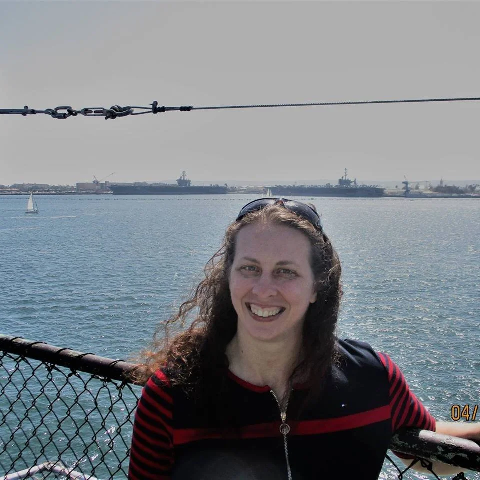 A smiling woman with long curly brown hair stands by the water with ships in the distance behind her.