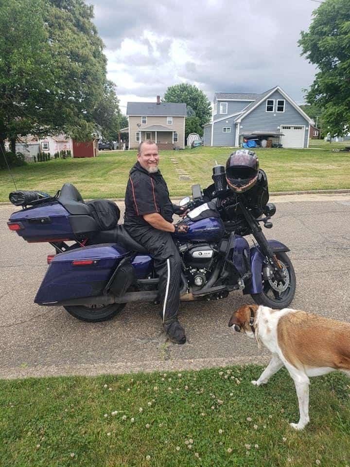 Motorcycle riding man sits on his bike with a dog coming up to him.