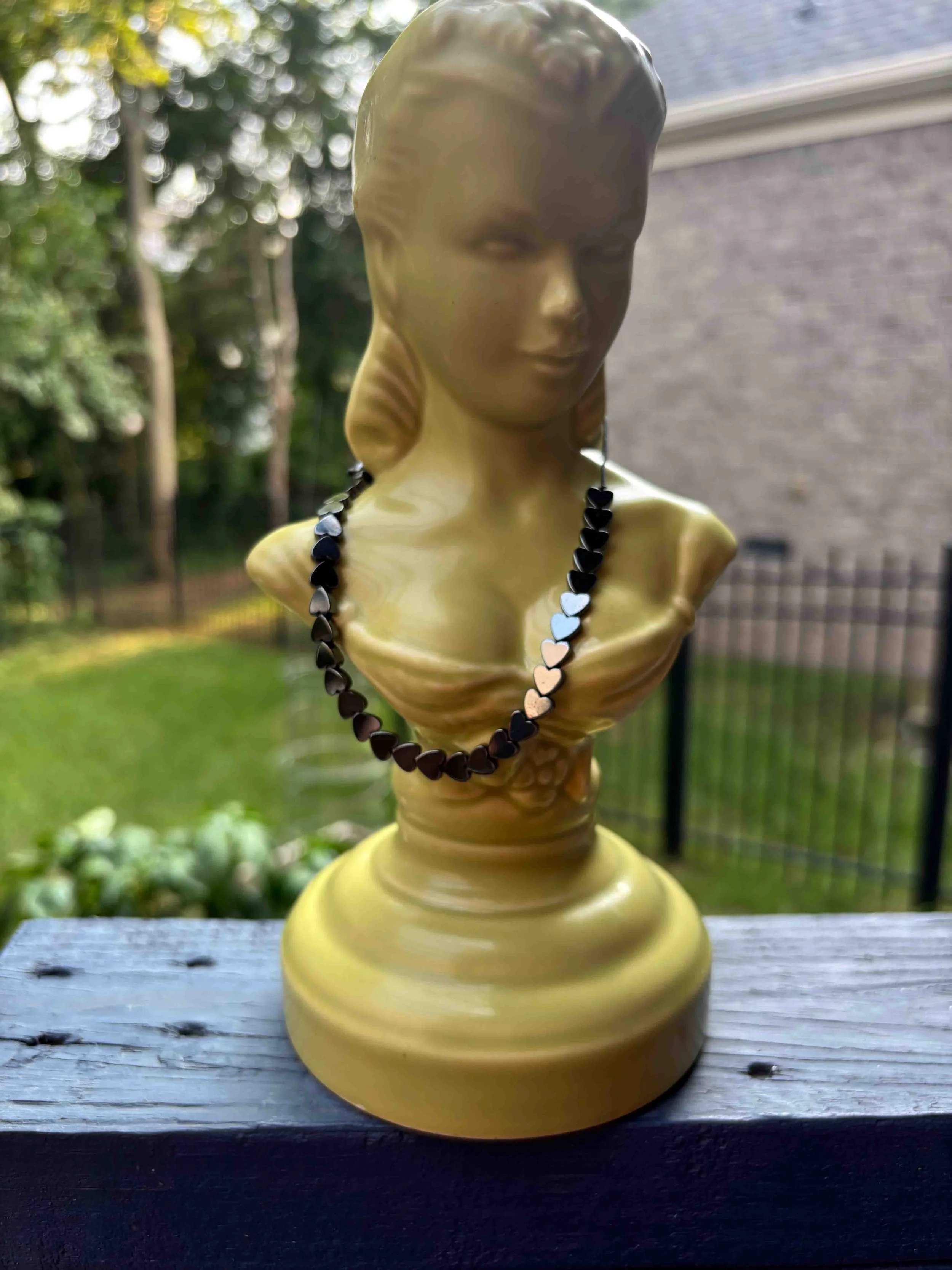 Heart-shaped hematite bead bracelet displayed on a mustard-yellow vintage ceramic bust outdoors.
