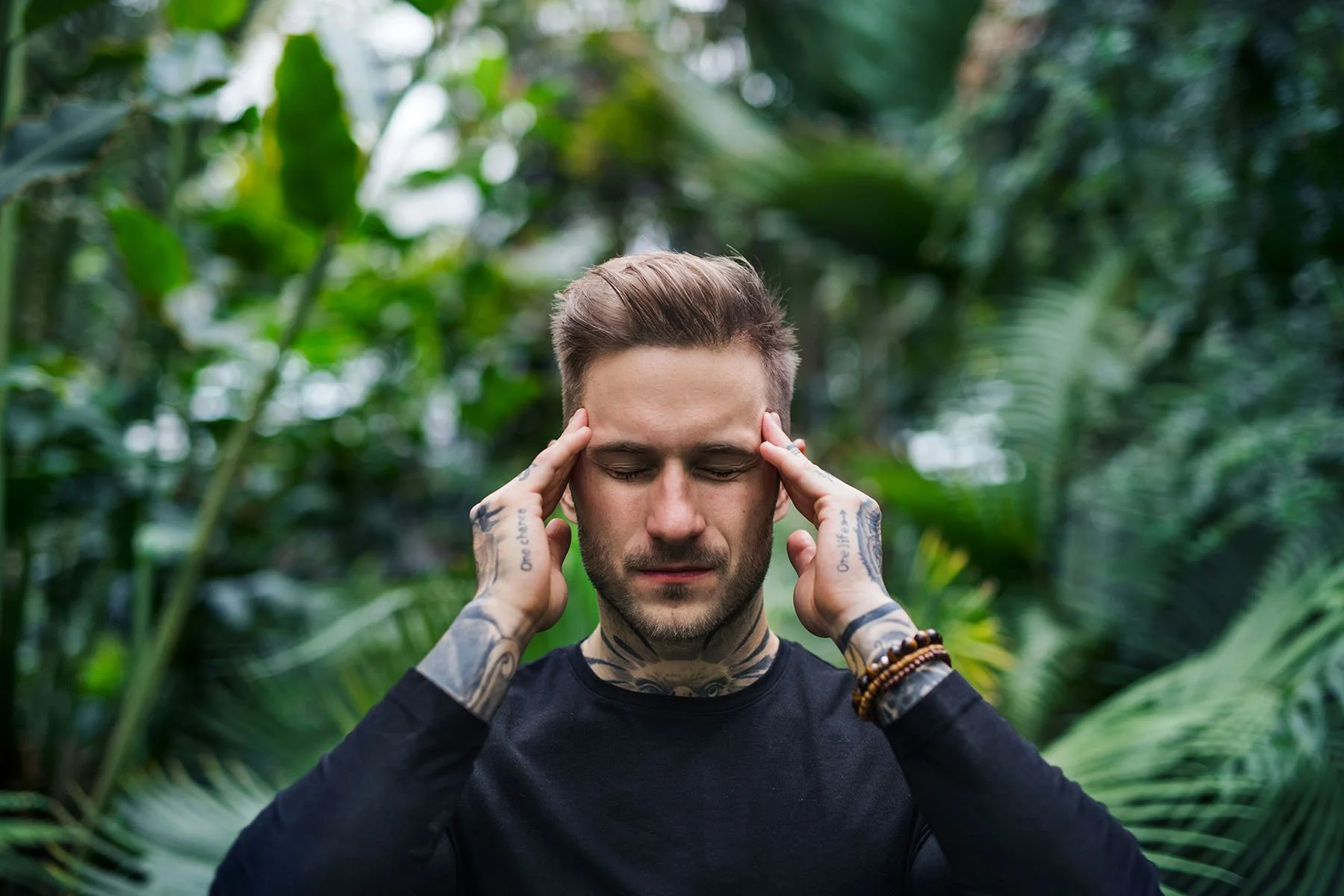 Person with eyes closed and hands resting on their temples while standing in a lush tropical setting.