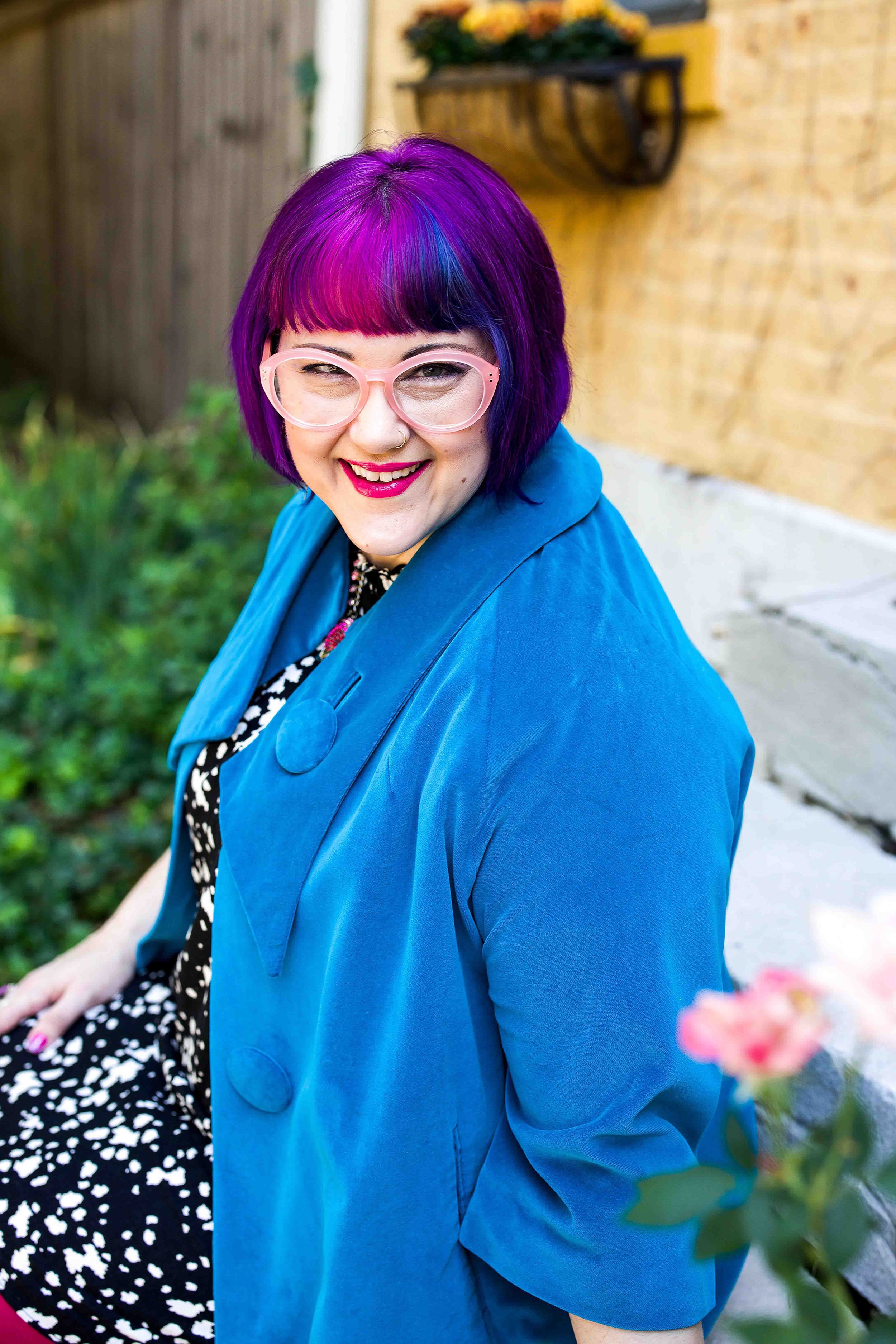 Outdoor portrait of a smiling person with vibrant purple hair and pink glasses wearing a blue coat, seated near a garden with flowers in the foreground.