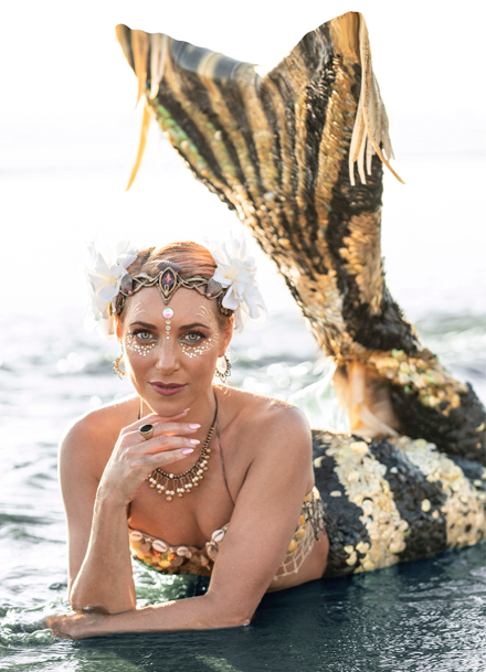 Mermaid performer resting in shallow ocean water wearing a shimmering gold-and-black mermaid tail with seashell-inspired jewelry and face gems.