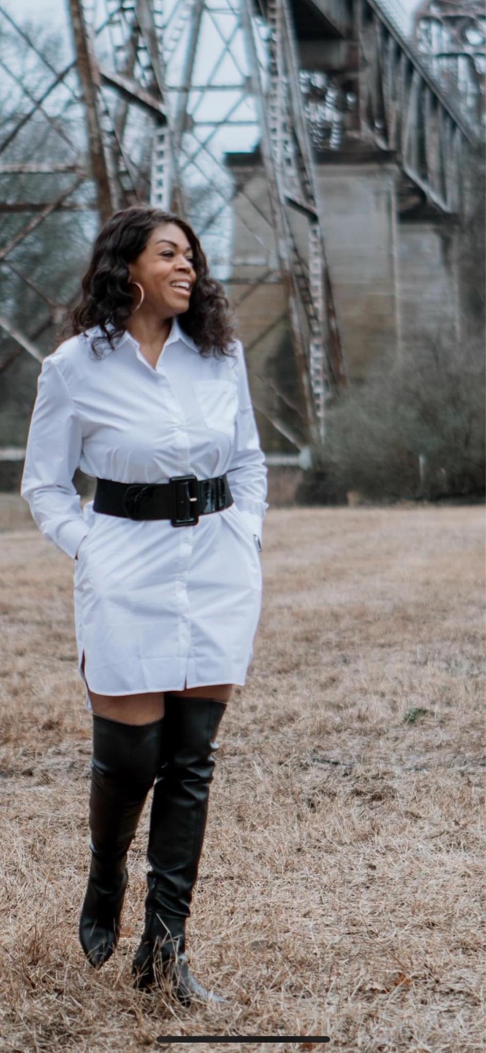 Black woman wearing a white shirt dress with a black belt and black thigh high boots stands outside, smiles and looks to the side