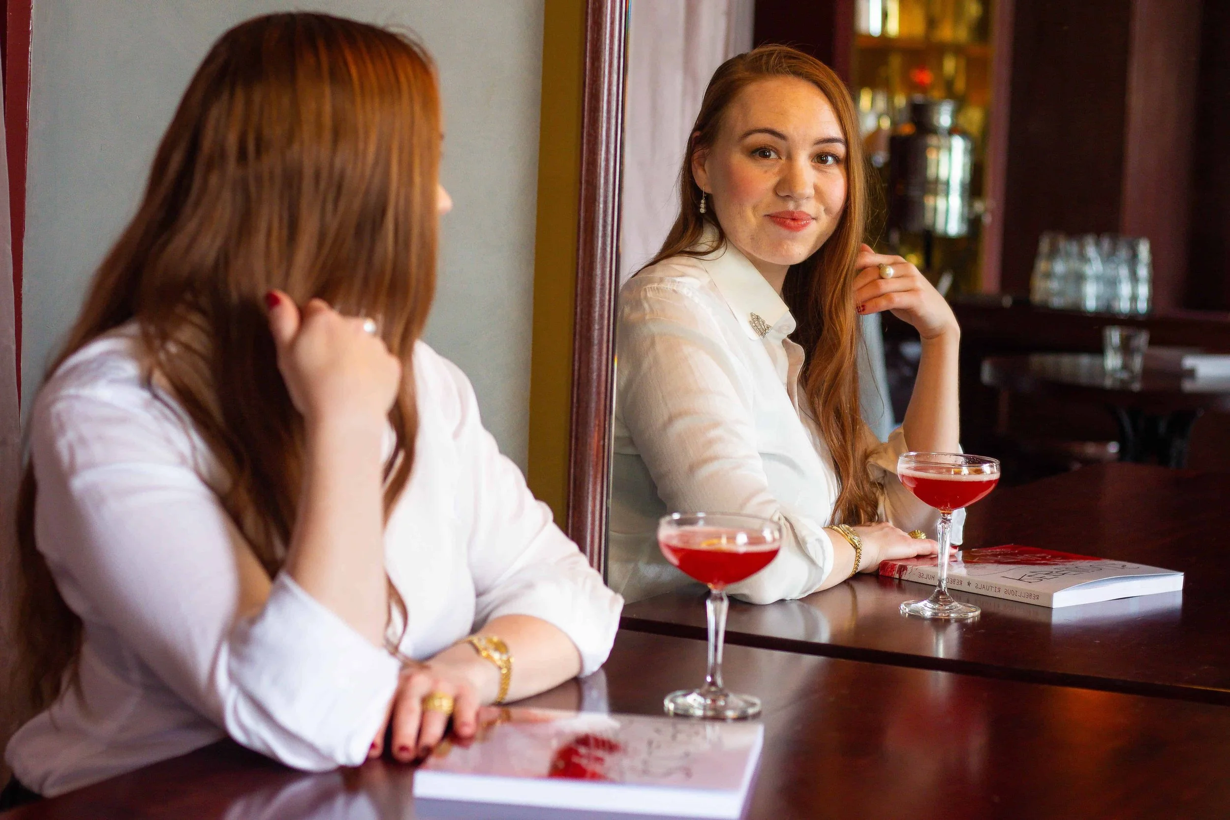 Author looking into a mirror with an open copy of Rebellious Rituals and a cocktail on the table.