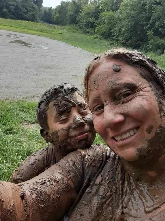 A woman and child smile for a muddy outdoor selfie near a grassy field and tree line.