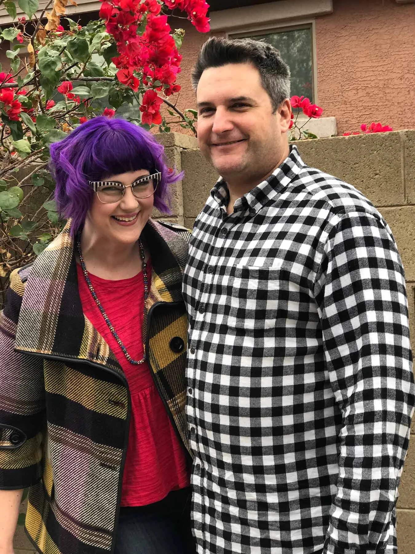 A smiling couple standing together outdoors in front of blooming red flowers, wearing casual clothes and sharing a joyful moment.