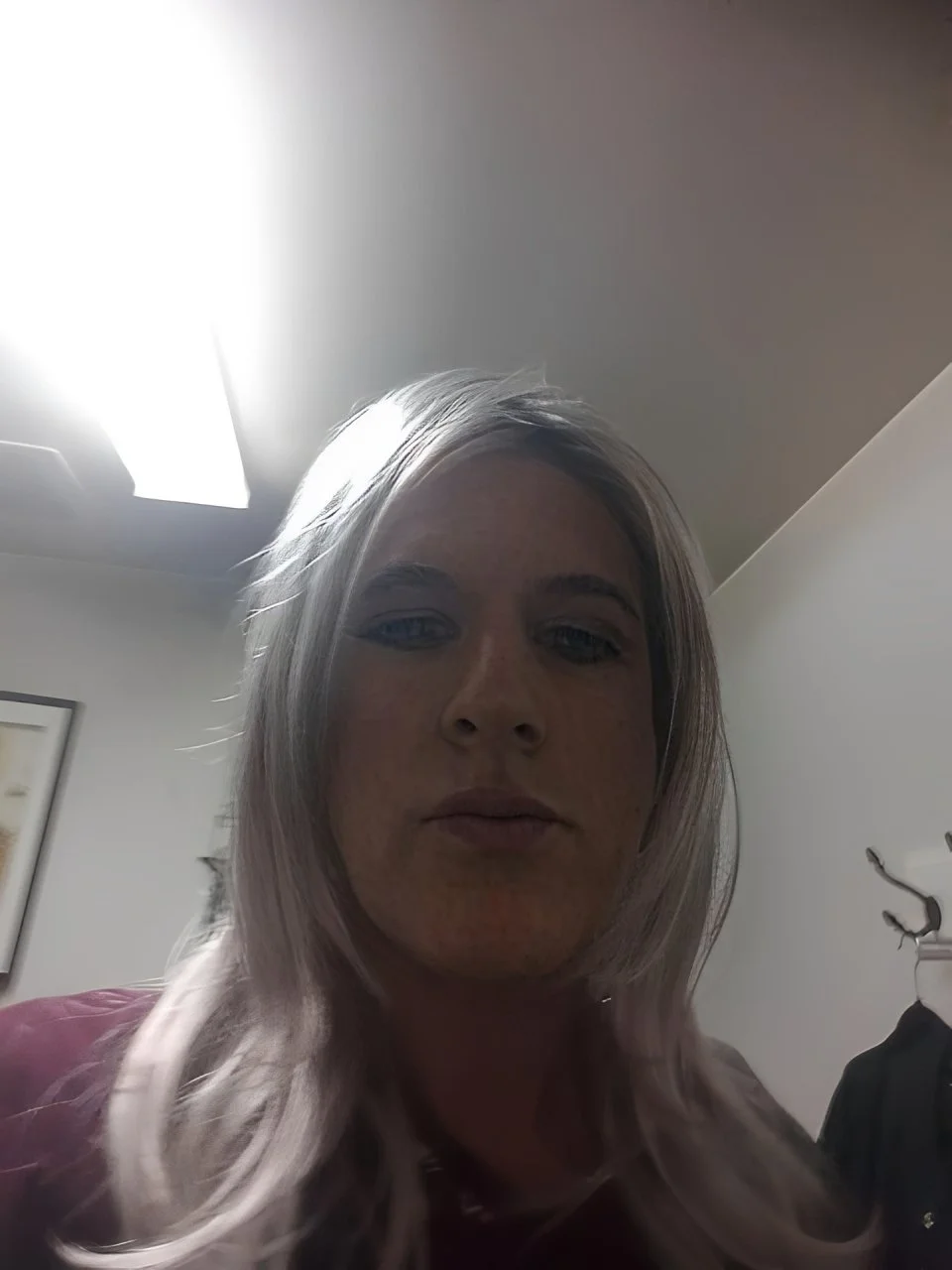 Low-light selfie of a person with long silver hair sitting indoors beneath bright overhead lighting, wearing subtle makeup and a burgundy top.