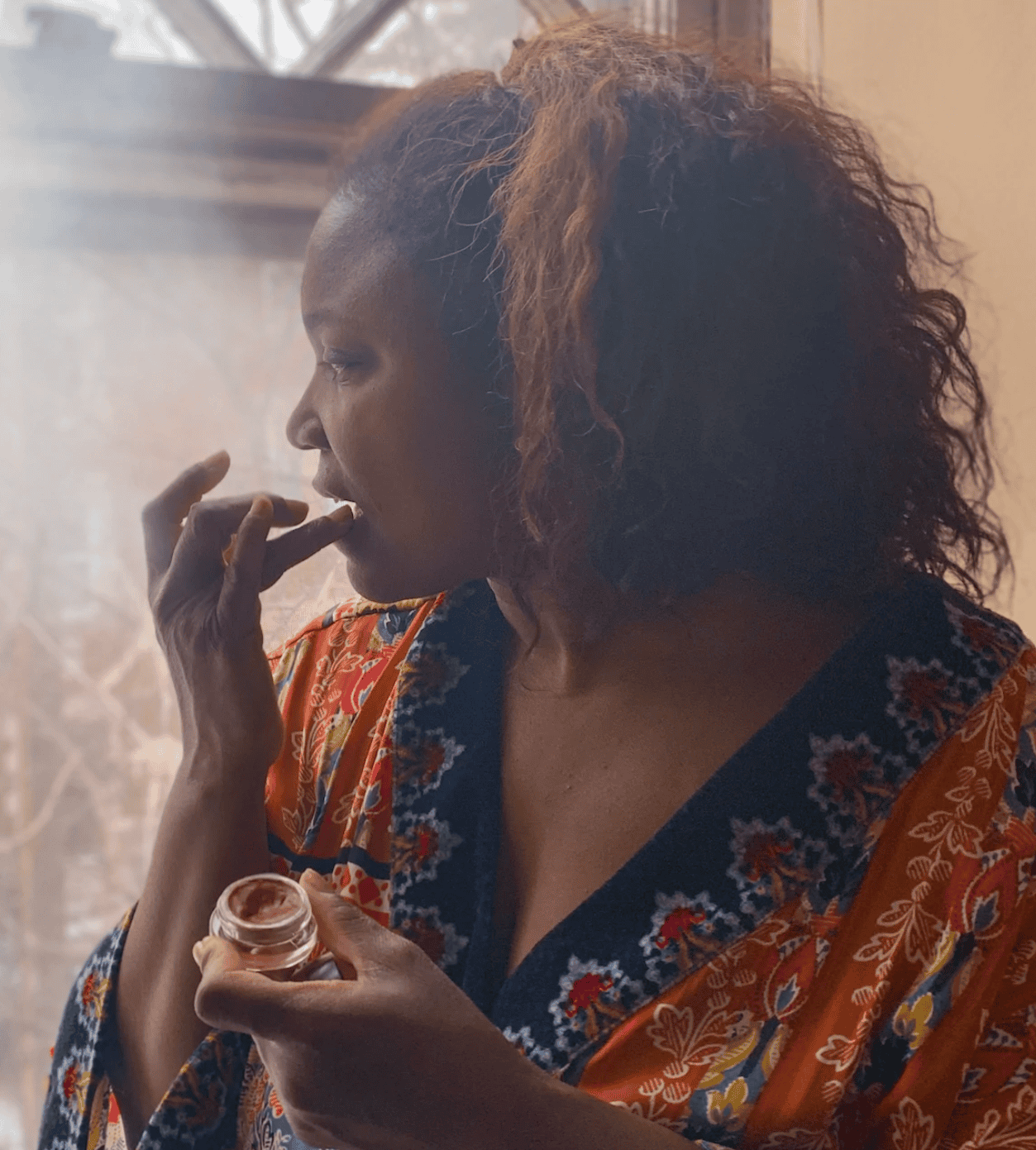 Woman applying lip gloss in front of a window.